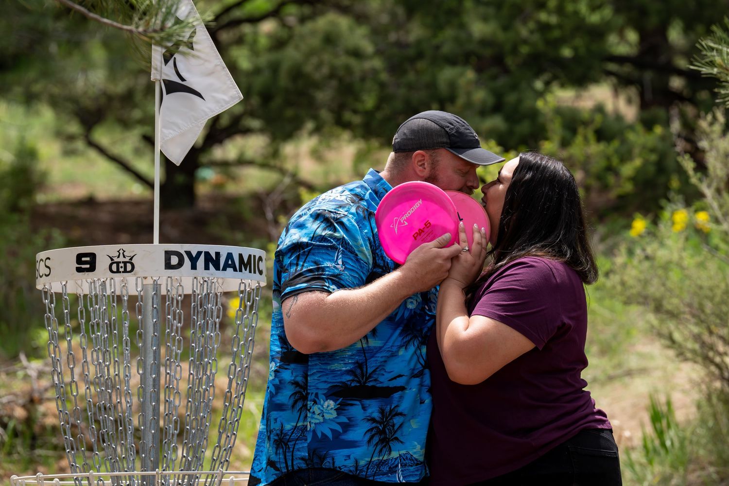 A couple sharing a tender moment near a disc golf basket in a mountain setting.