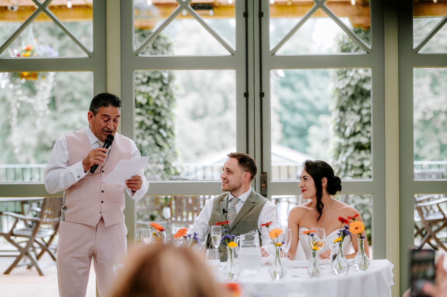 Wedding reception guests enjoying speeches in bright conservatory setting.