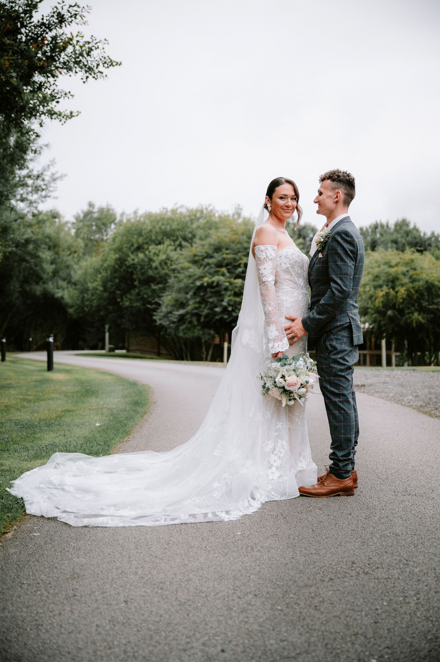 A romantic wedding couple poses on a winding path surrounded by lush greenery and trees.
