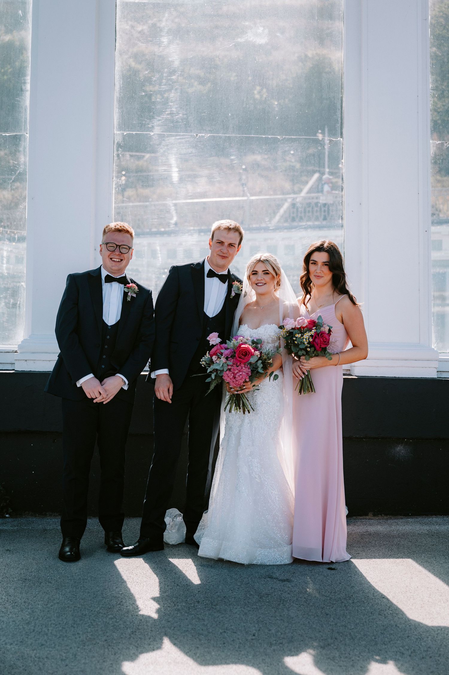 Wedding party members pose together in formal attire against bright window backdrop.