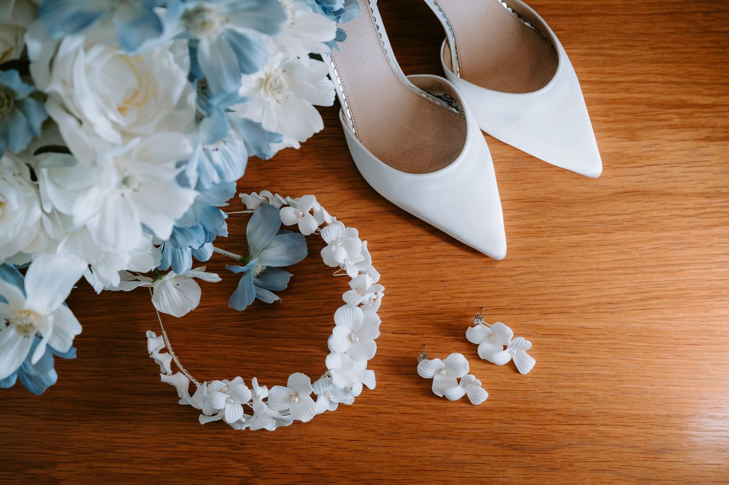 Wedding accessories featuring white heels, blue flowers and pearl hair accessories on wooden surface.