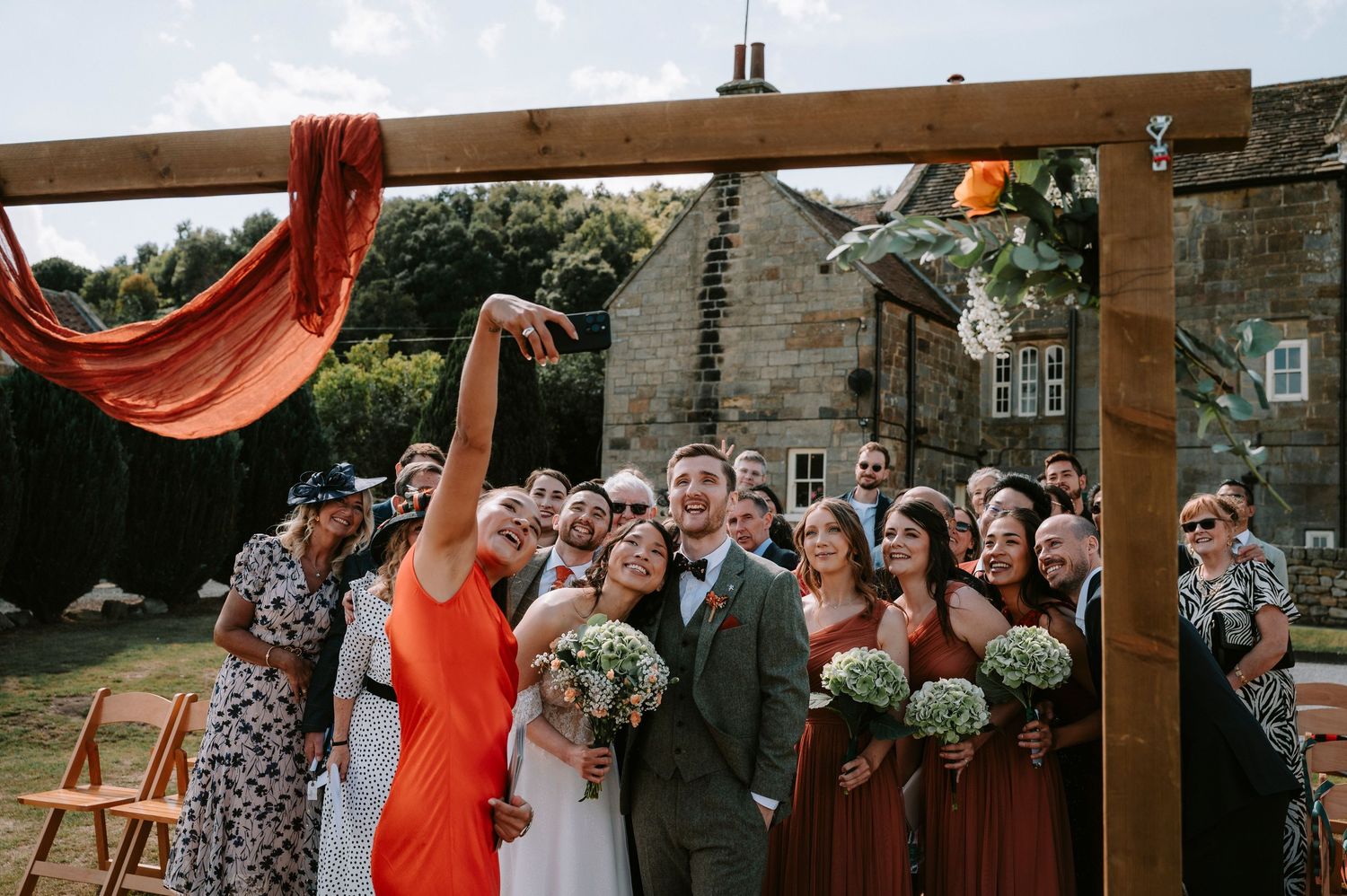 Wedding celebration under wooden pergola decorated with flowing red fabric and floral arrangements.