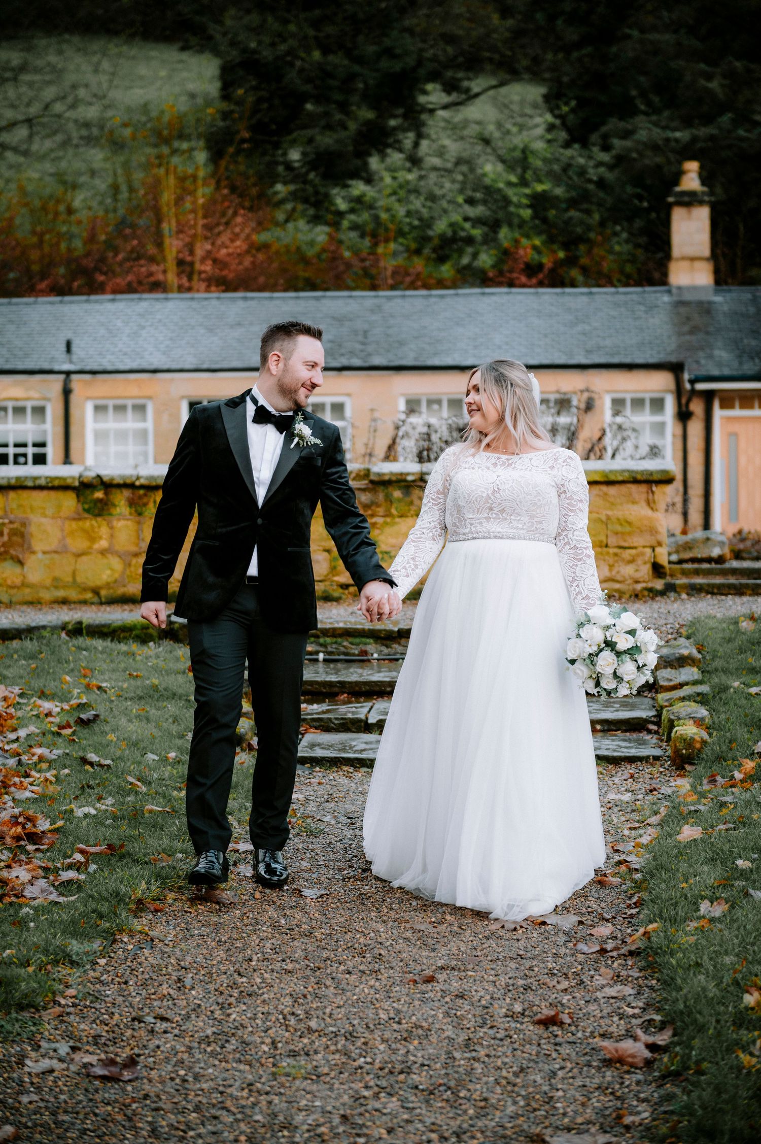 A couple walks hand in hand on a gravel path in front of a charming stone cottage.