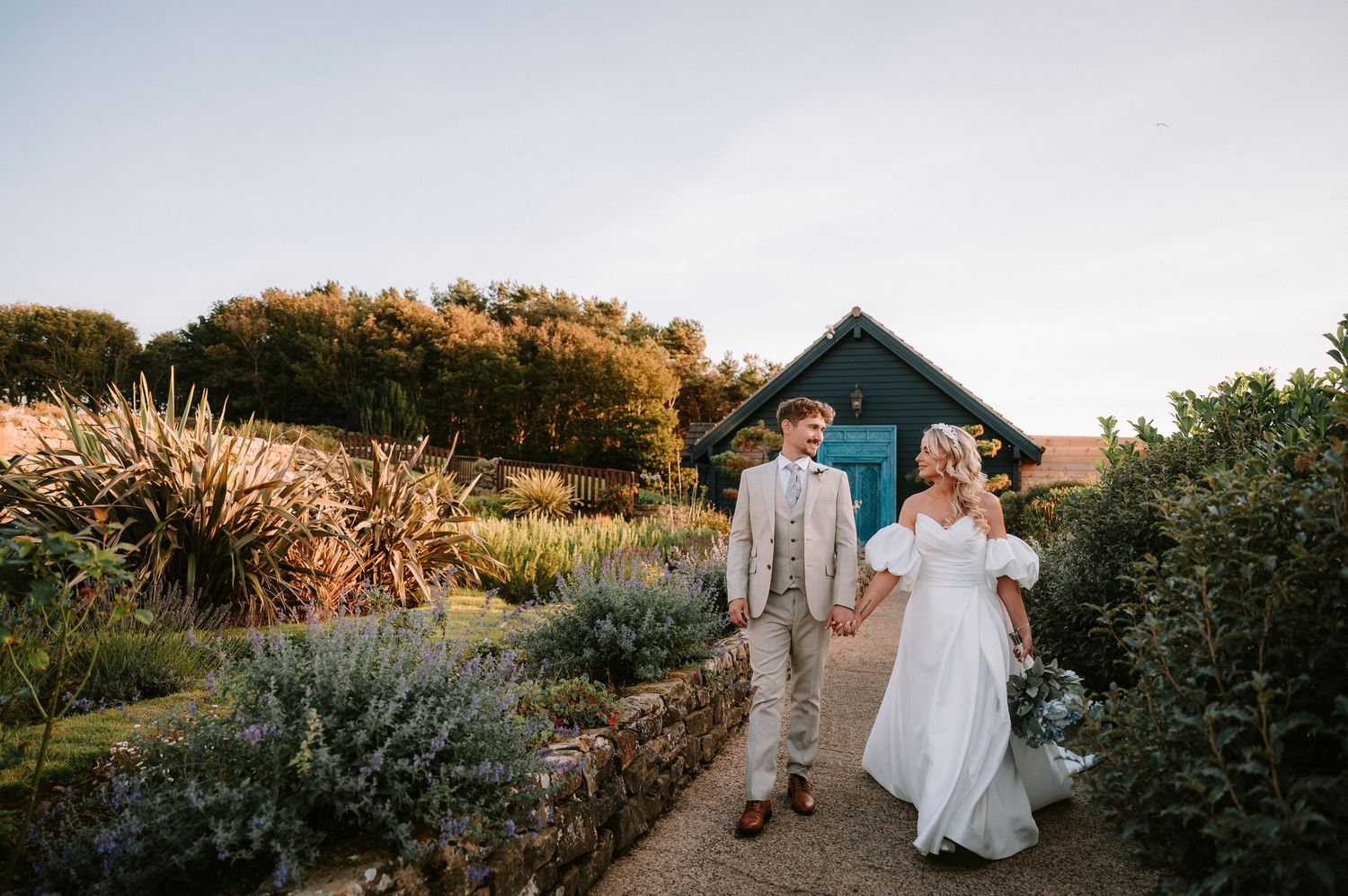 Couple walking along garden path with rustic barn and autumn foliage in background at sunset.