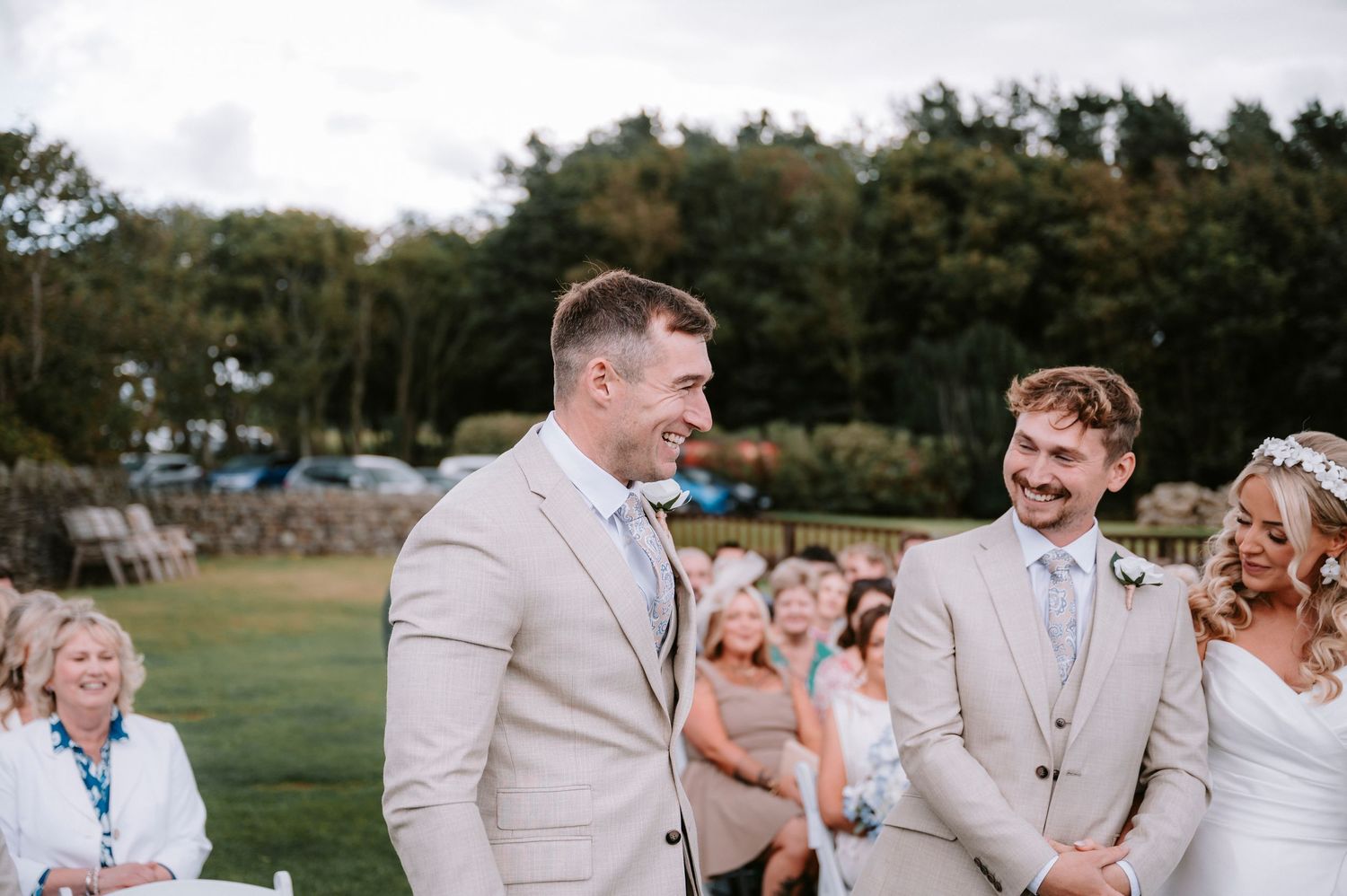 Wedding party laughs together outdoors on a sunny day wearing light colored formal attire.