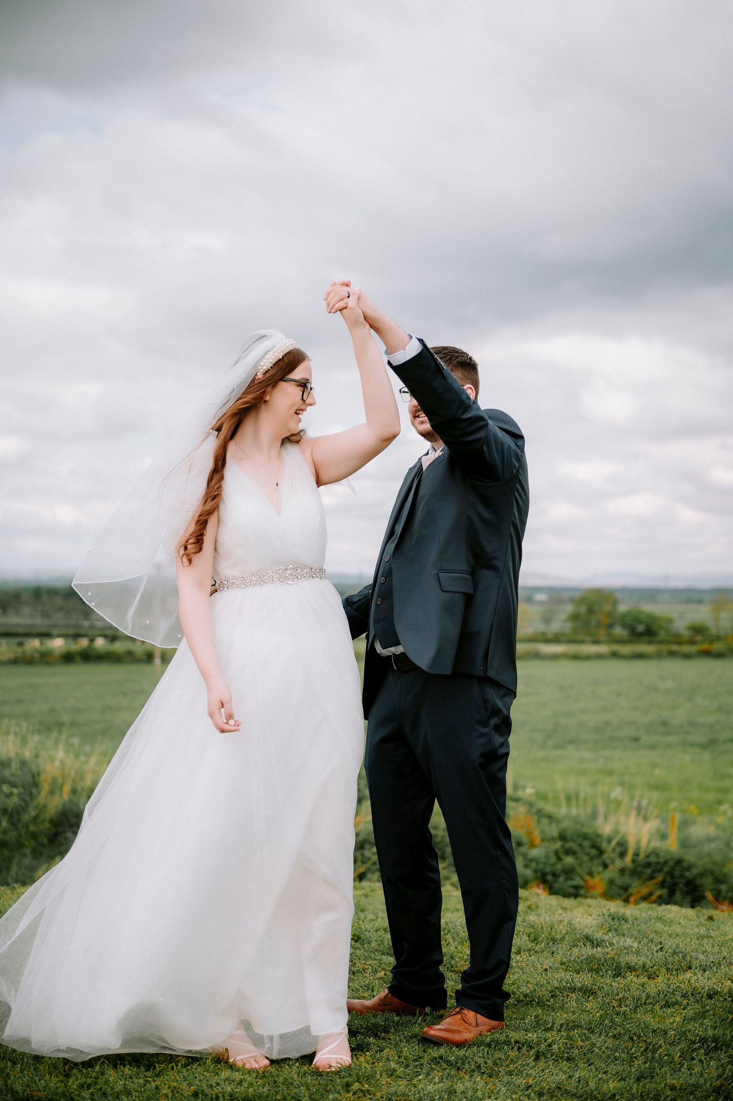 Couple dancing joyfully on a hilltop with dramatic cloudy sky backdrop.