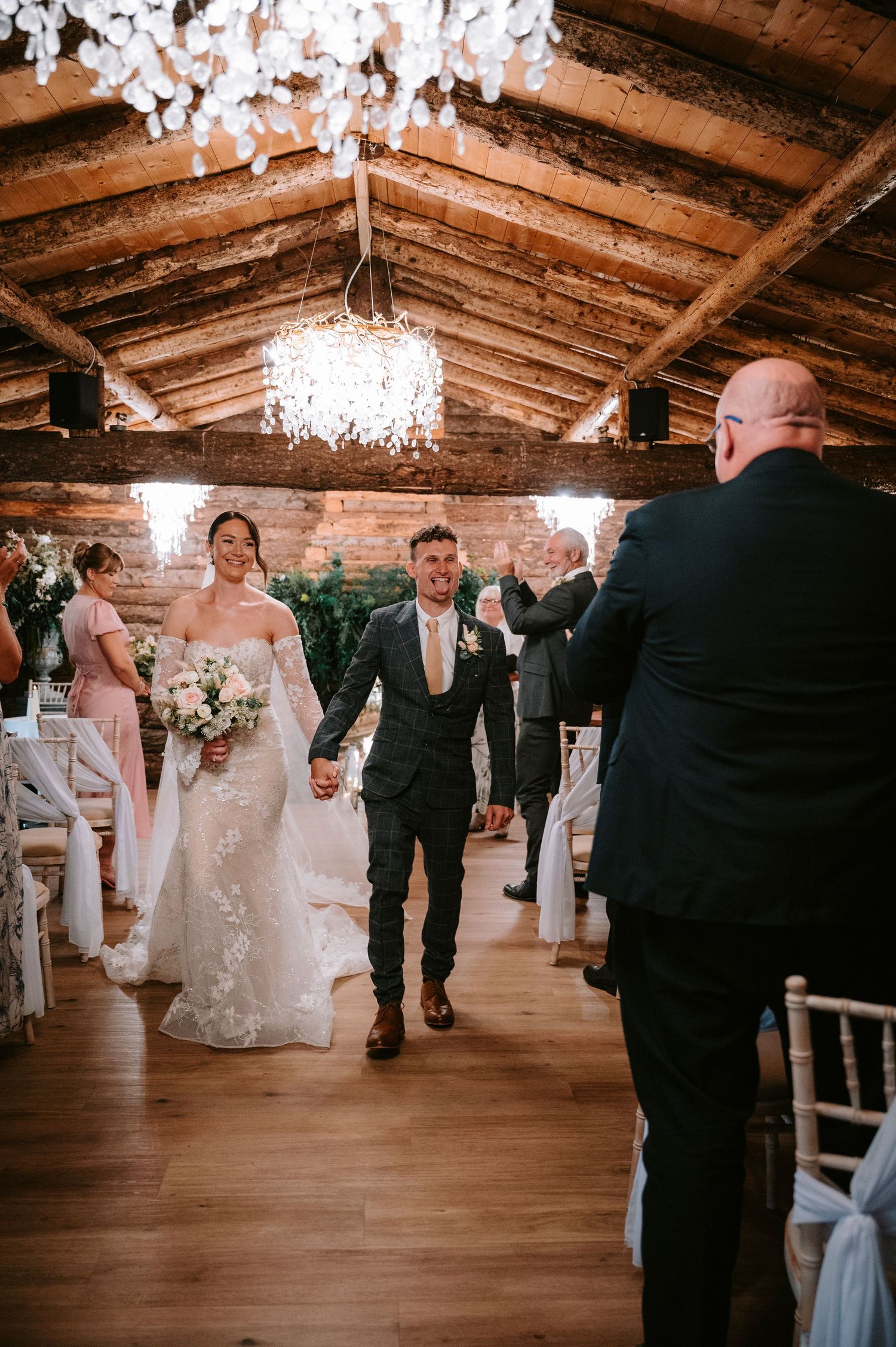 Wedding couple exits ceremony under crystal chandelier in rustic wooden beam venue.