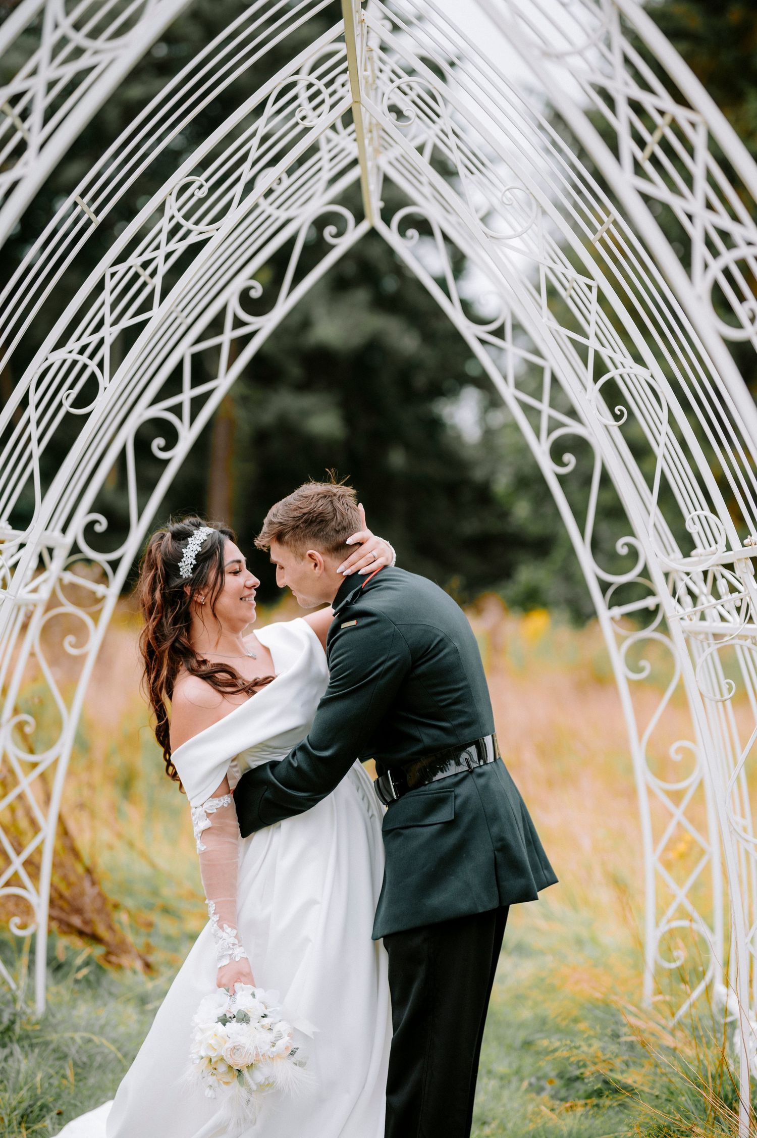 A wedding couple embraces under an elegant white metal arch in a natural outdoor setting.