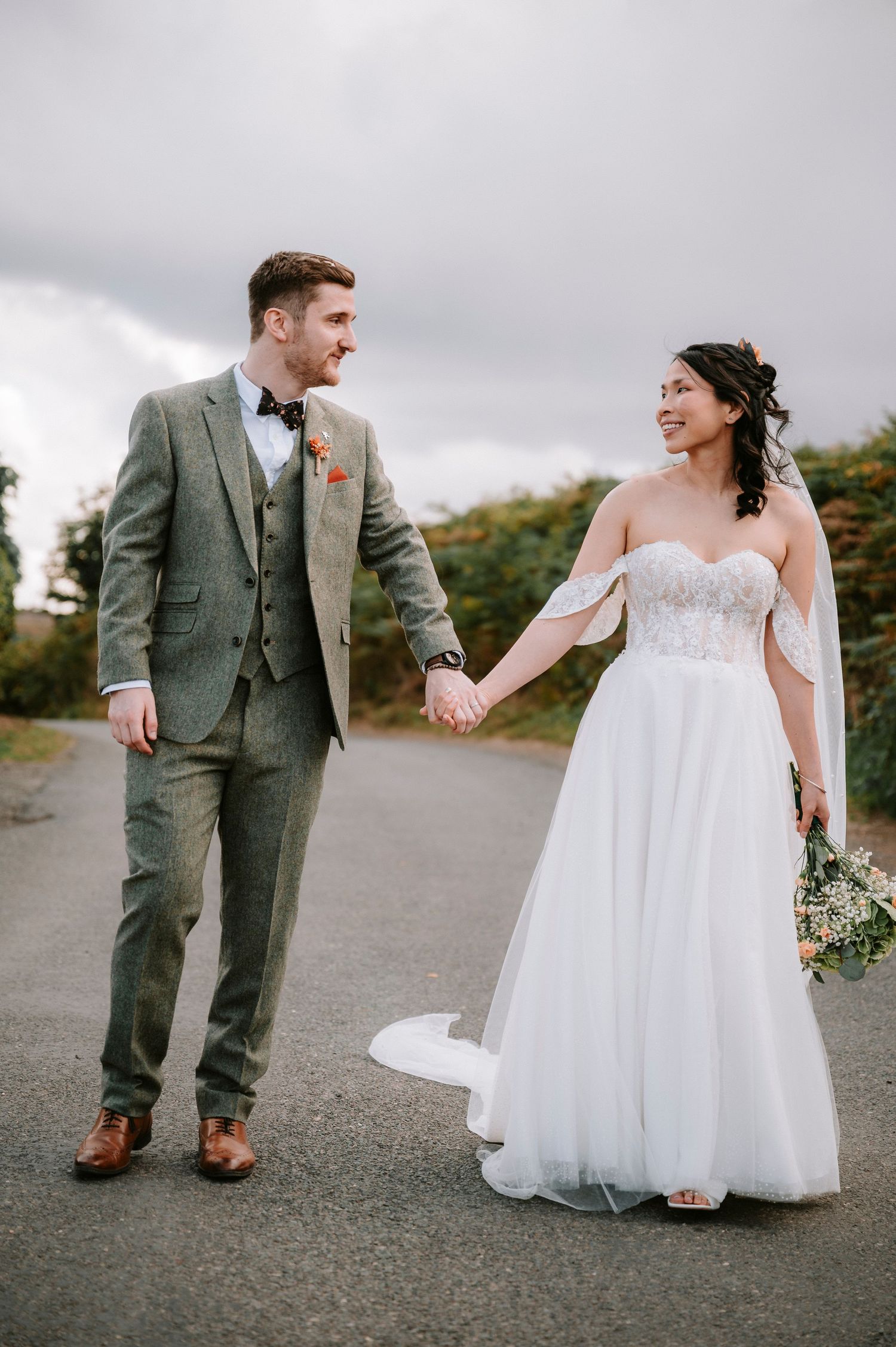Couple in wedding attire walking hand in hand on a country road with cloudy sky backdrop.