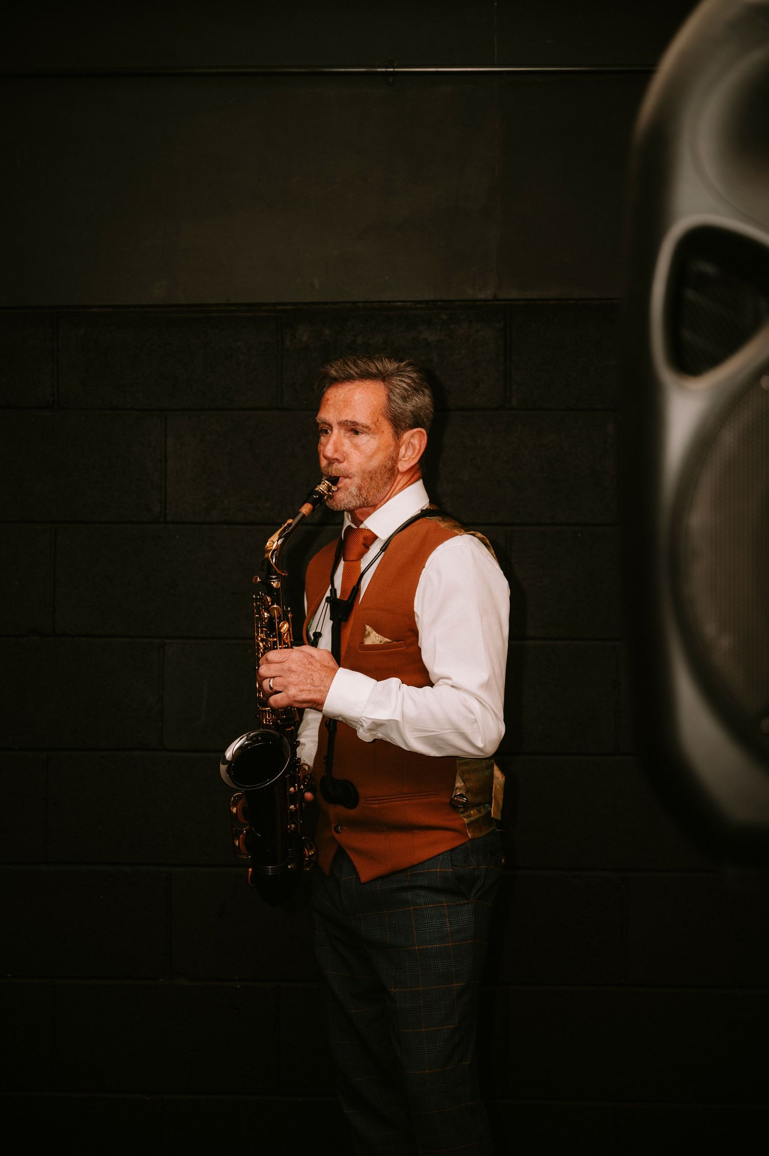A musician performs on saxophone wearing a brown vest and white shirt against a dark background.
