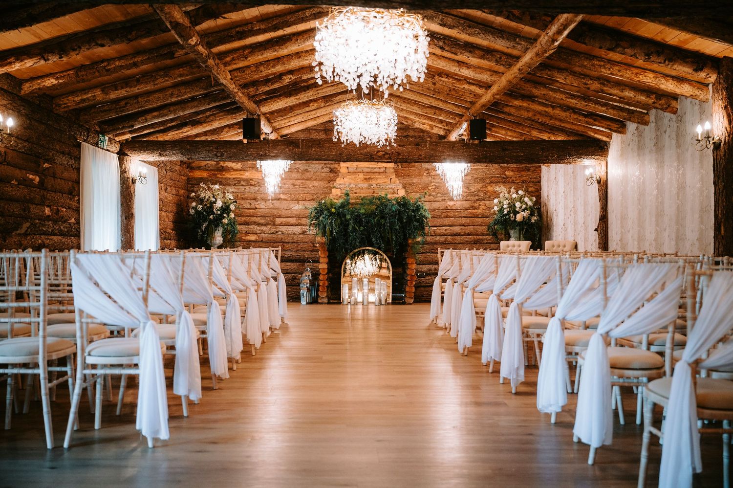 Rustic log cabin wedding venue with white chairs and crystal chandeliers set up for an indoor ceremony.