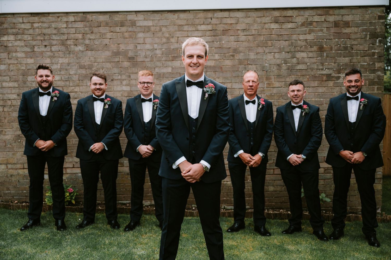 Wedding party in black tuxedos lined up against brick wall for formal photo.
