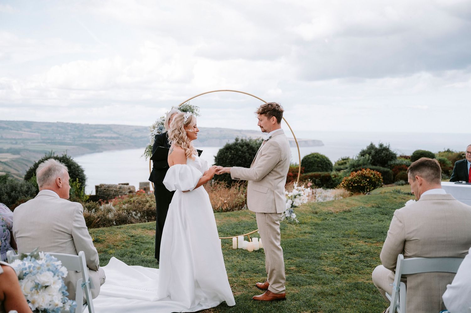 Romantic outdoor wedding ceremony with guests seated on a hillside overlooking a scenic mountain landscape.