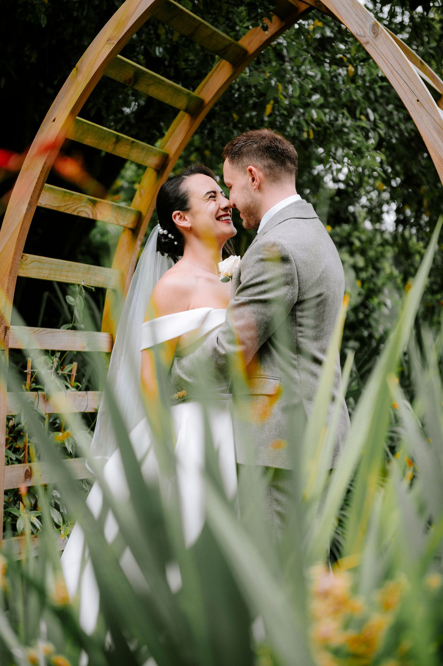Intimate wedding moment captured through garden foliage under wooden arch.
