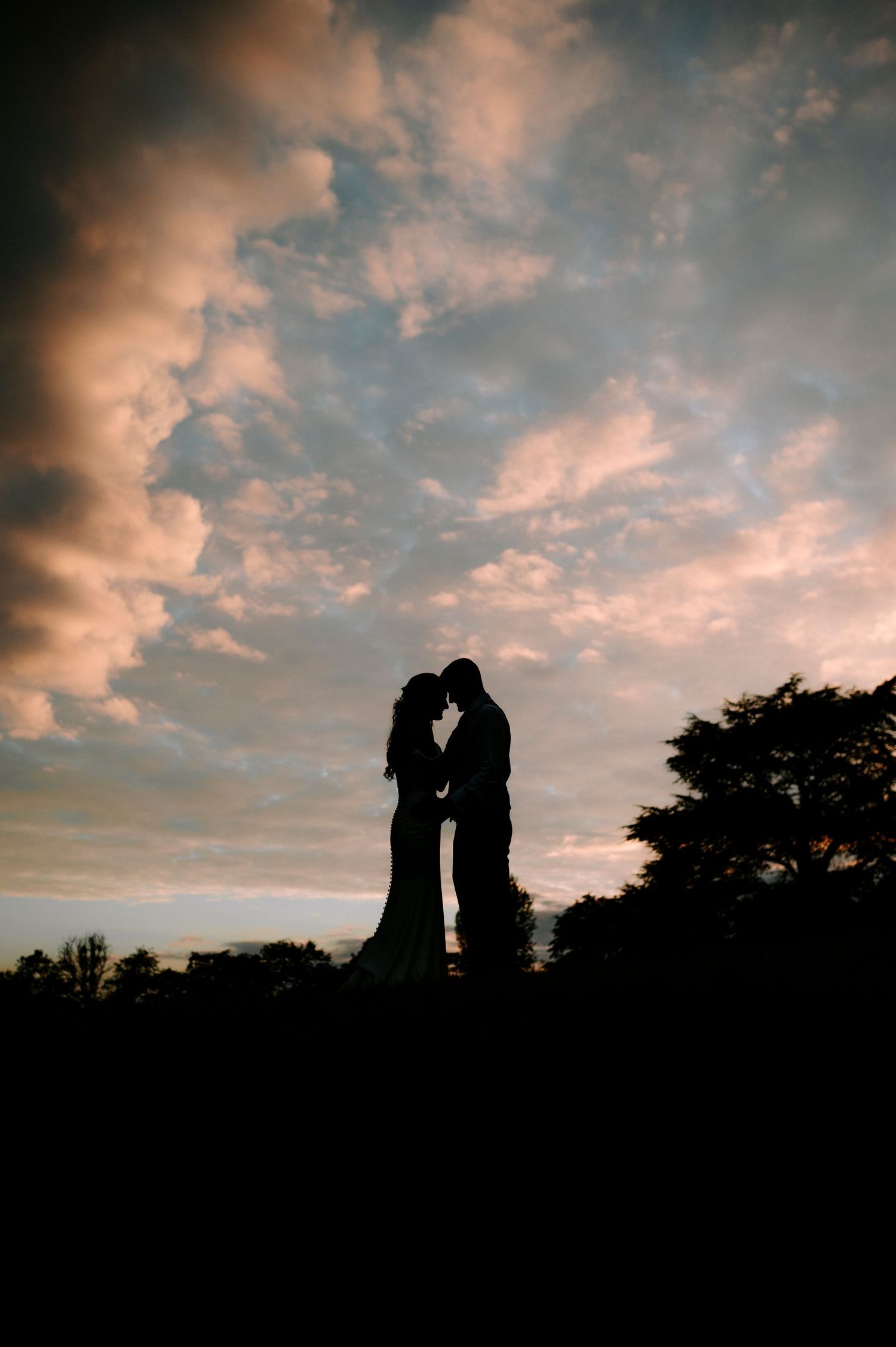 A romantic silhouette of a couple standing close together against a dramatic sunset sky with pink clouds.