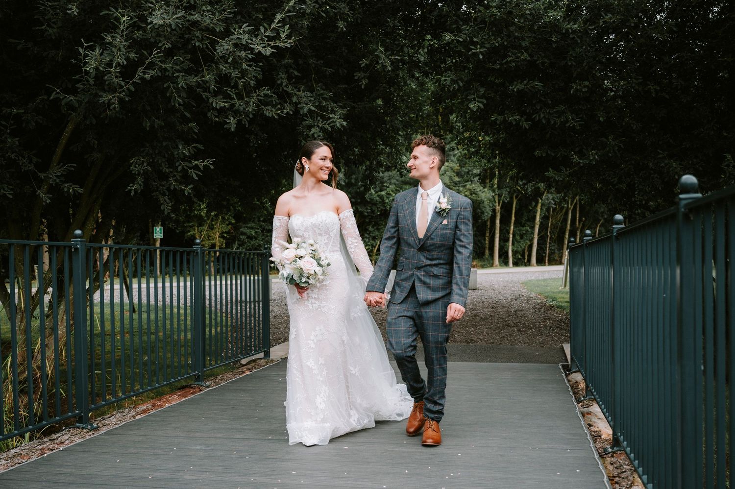 Couple walks hand in hand along a tree-lined path with iron fencing on a wedding day.