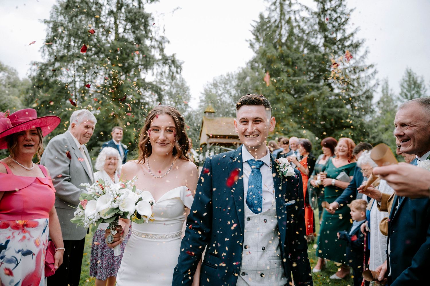 Joyful wedding celebration with guests throwing confetti in an outdoor garden setting.