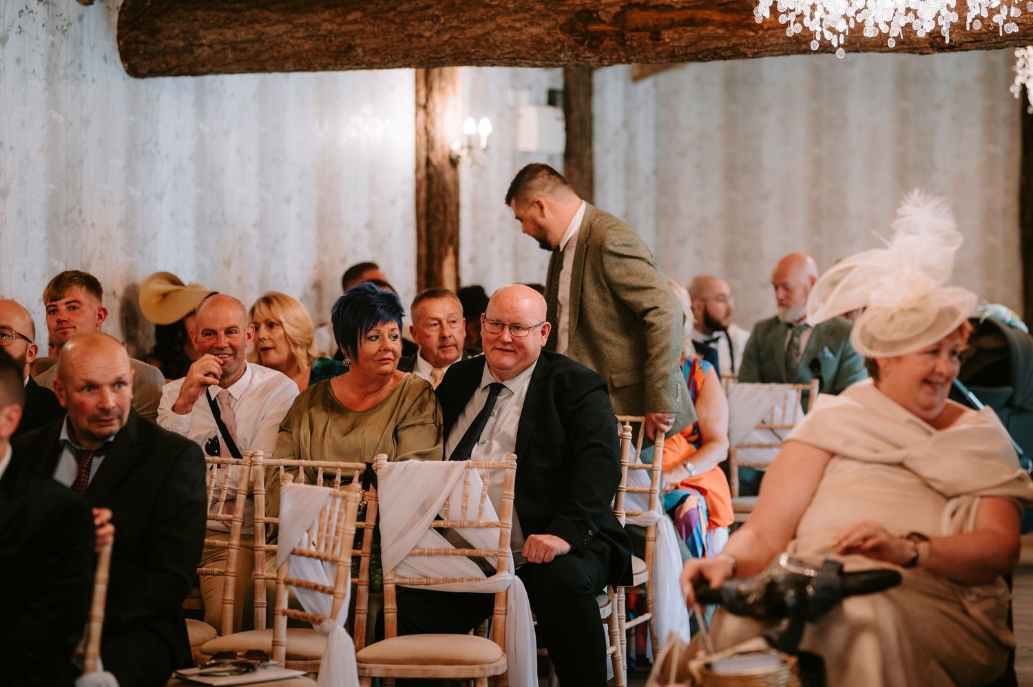Wedding reception guests seated in elegant chairs under warm chandelier lighting.