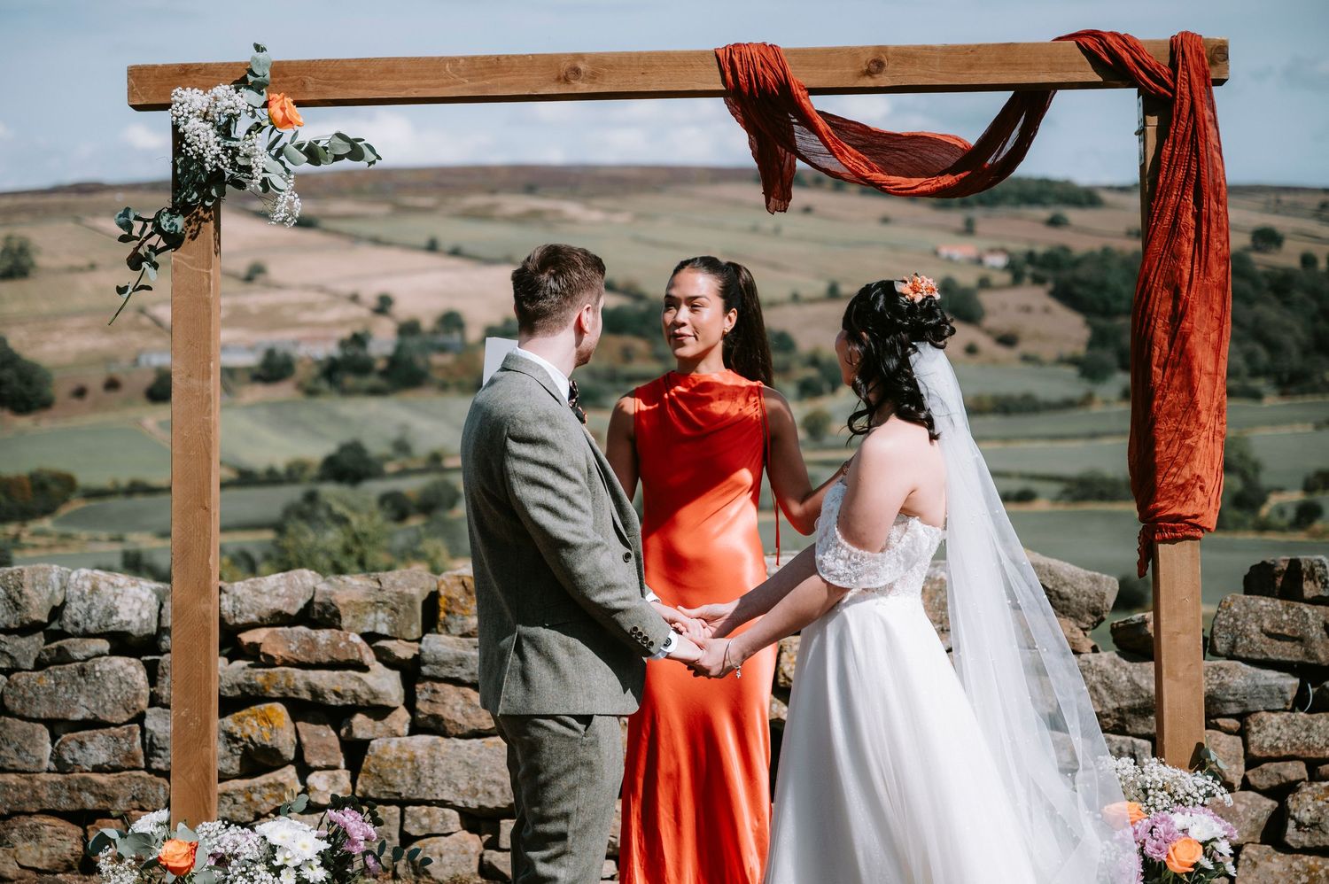 Outdoor wedding ceremony under wooden arch with red fabric draping against stone wall and rolling hills landscape.
