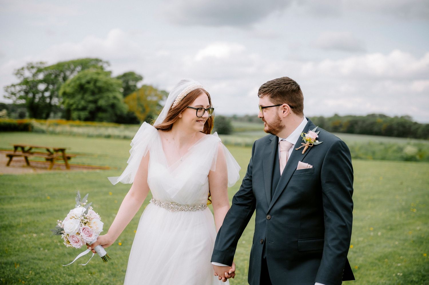 A couple poses together in an outdoor wedding photoshoot with rolling green fields in the background.