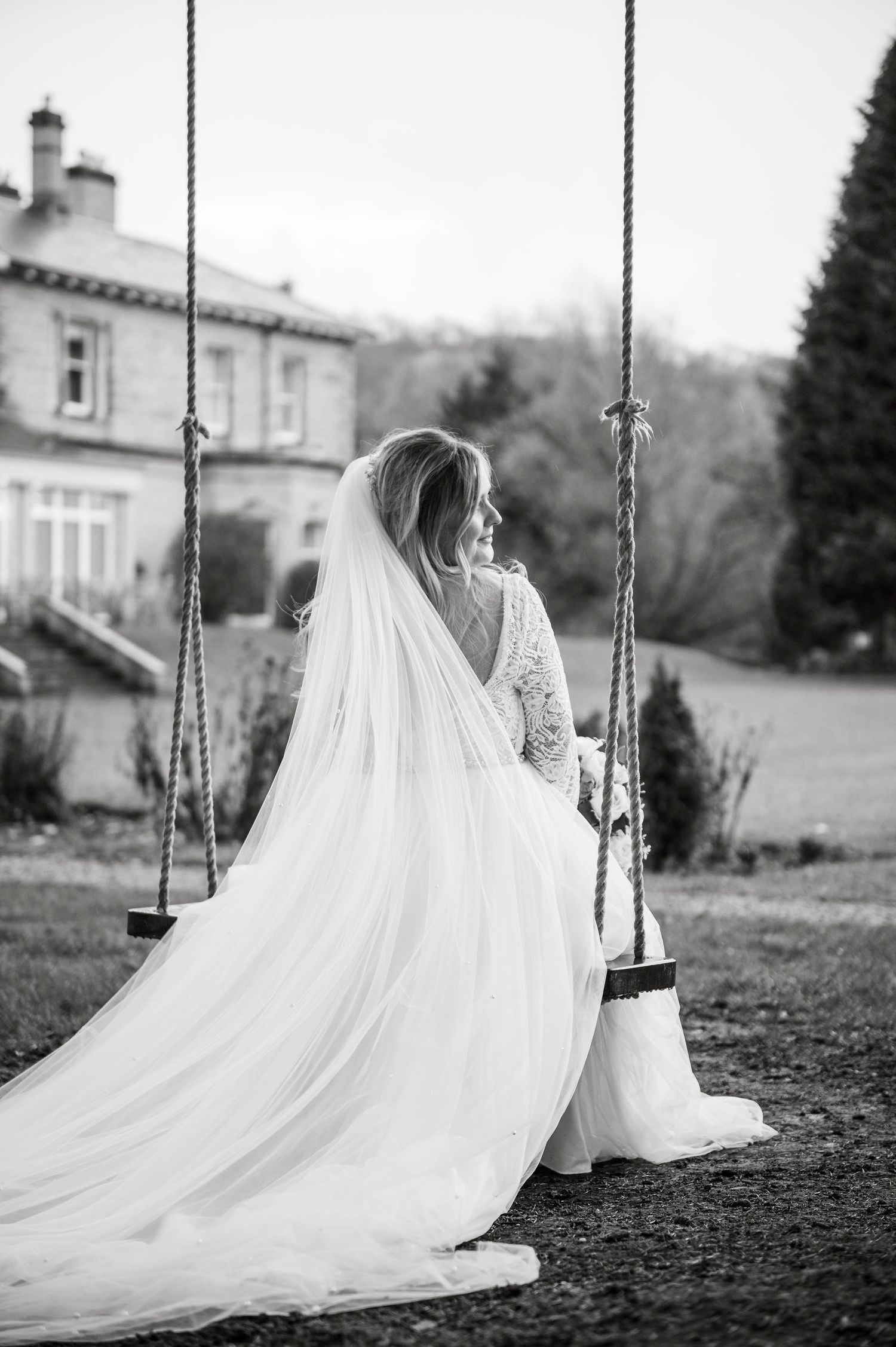 A bride in a flowing lace gown sits peacefully on a garden swing with a long veil trailing behind.