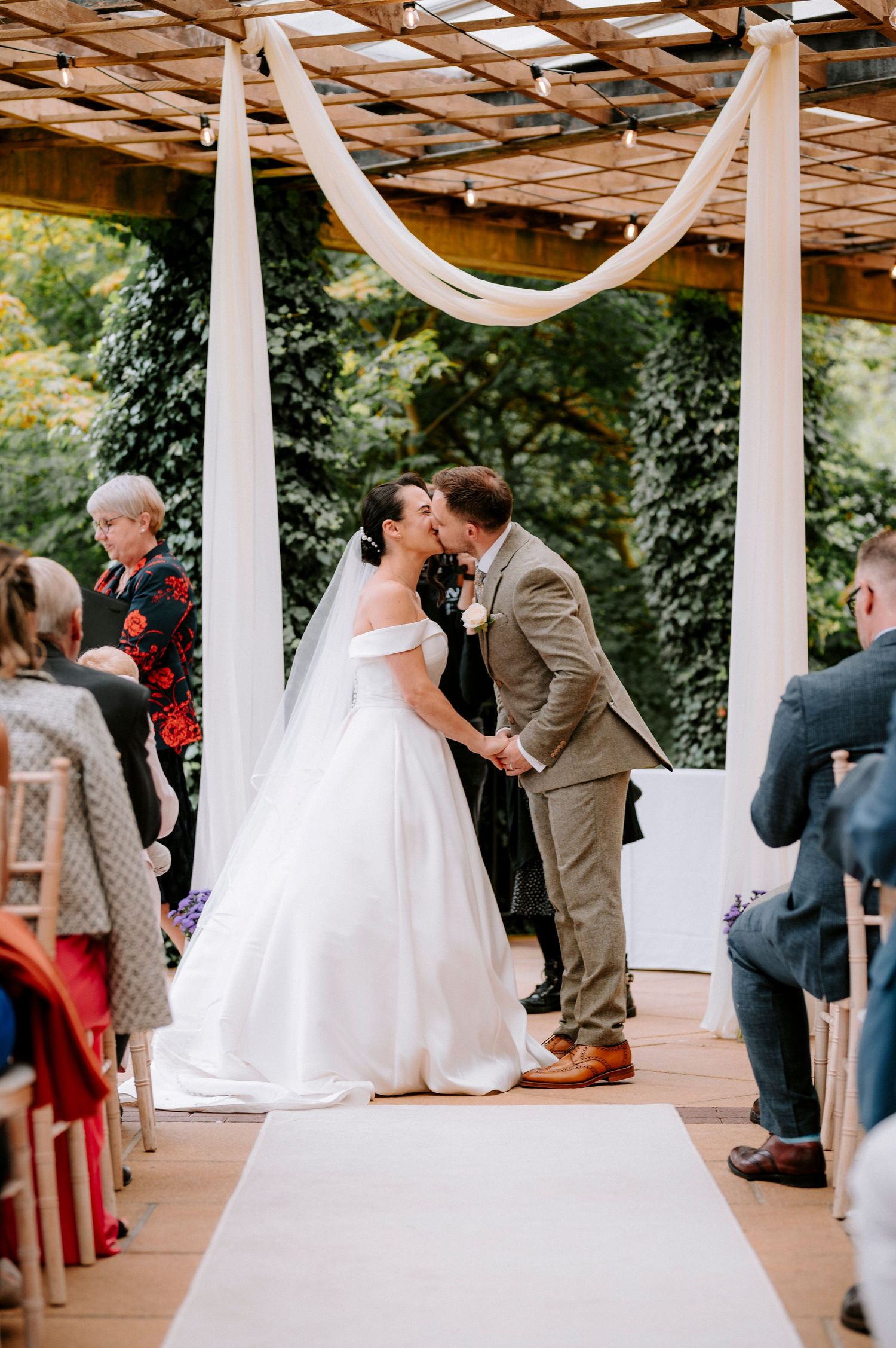 Wedding ceremony under wooden pergola with white fabric draping.