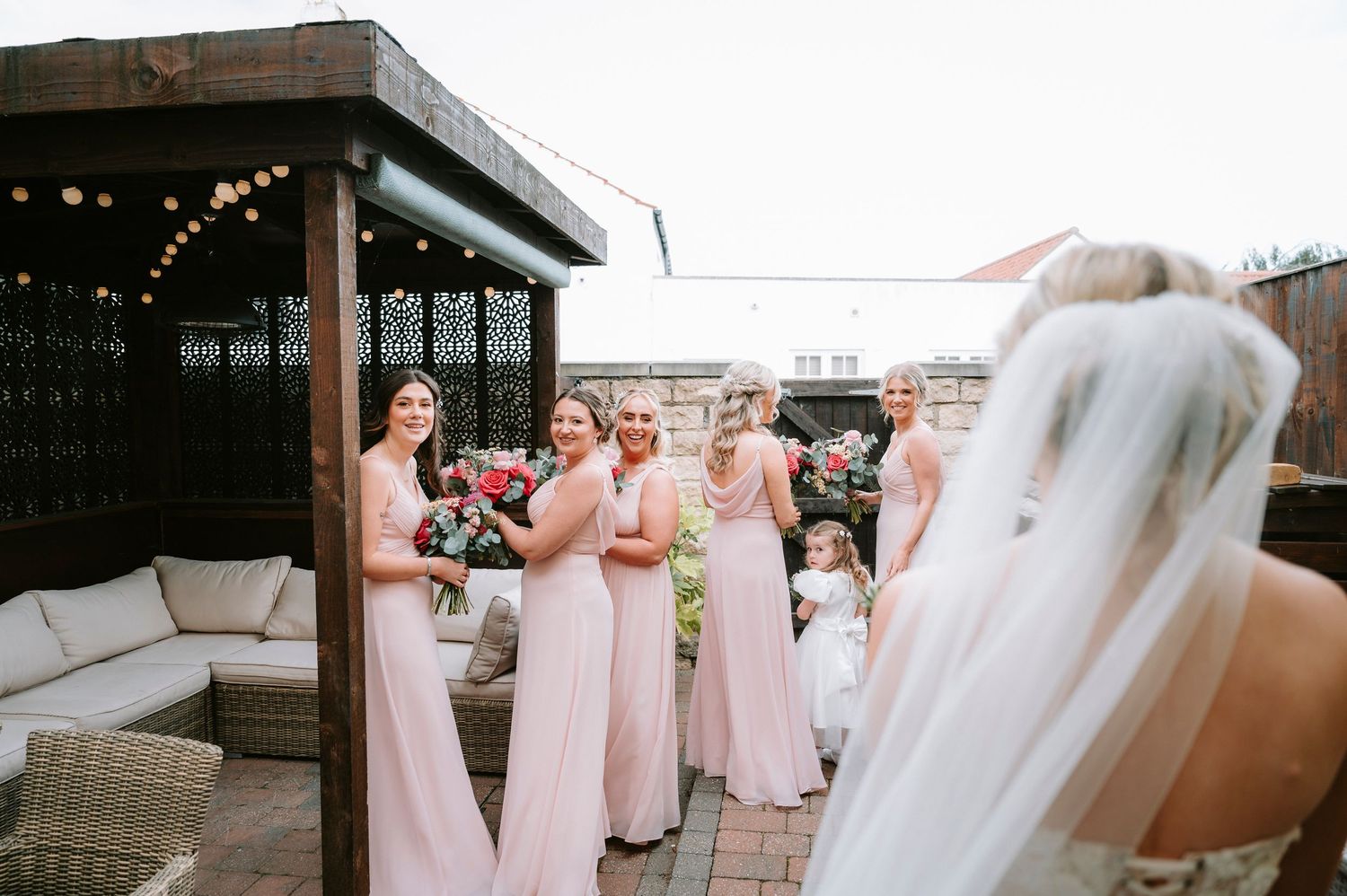 Wedding party in soft pink dresses gathered outdoors under string lights.