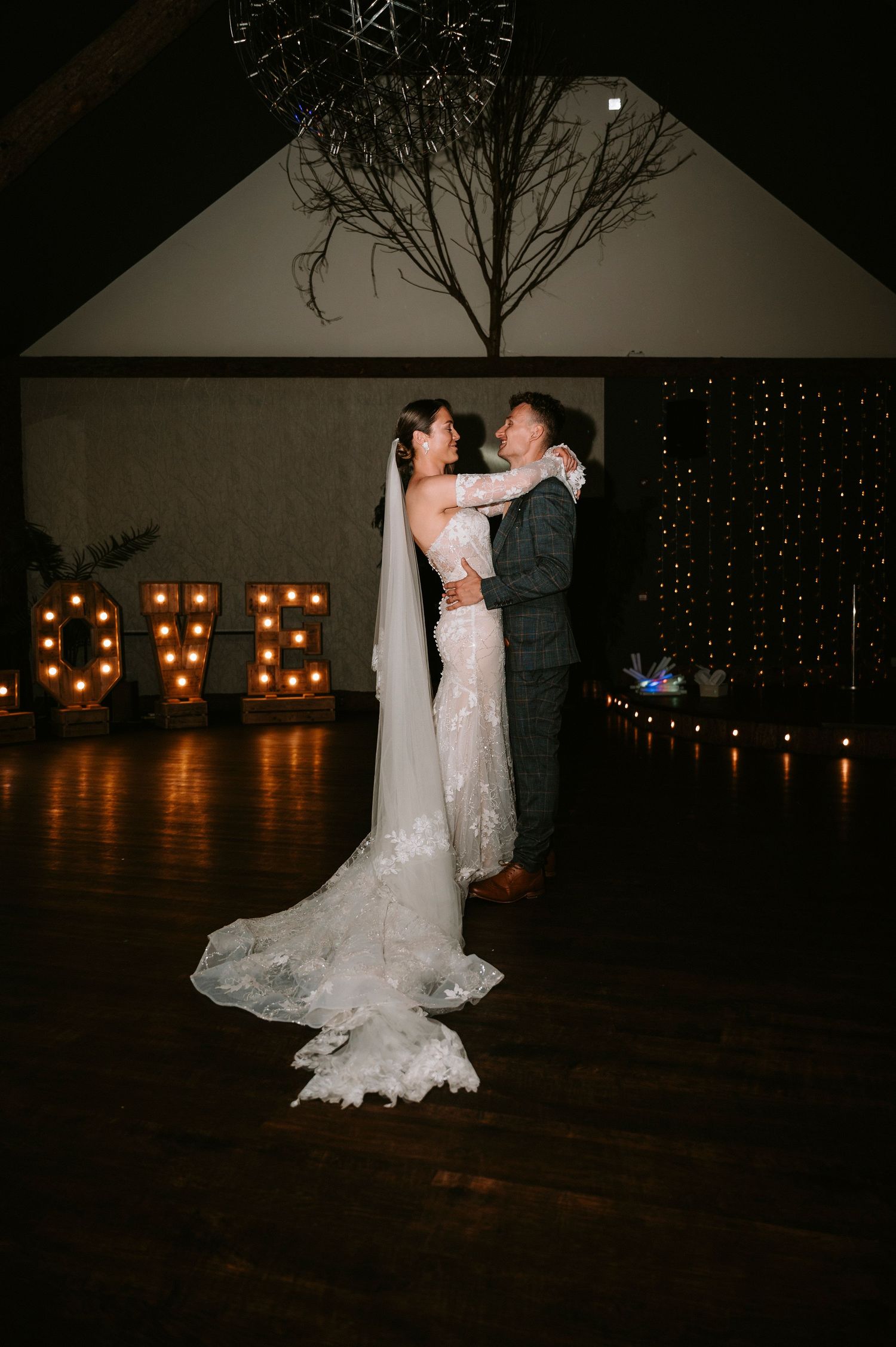 A couple shares their first dance in a dark reception venue with illuminated LOVE sign in background.