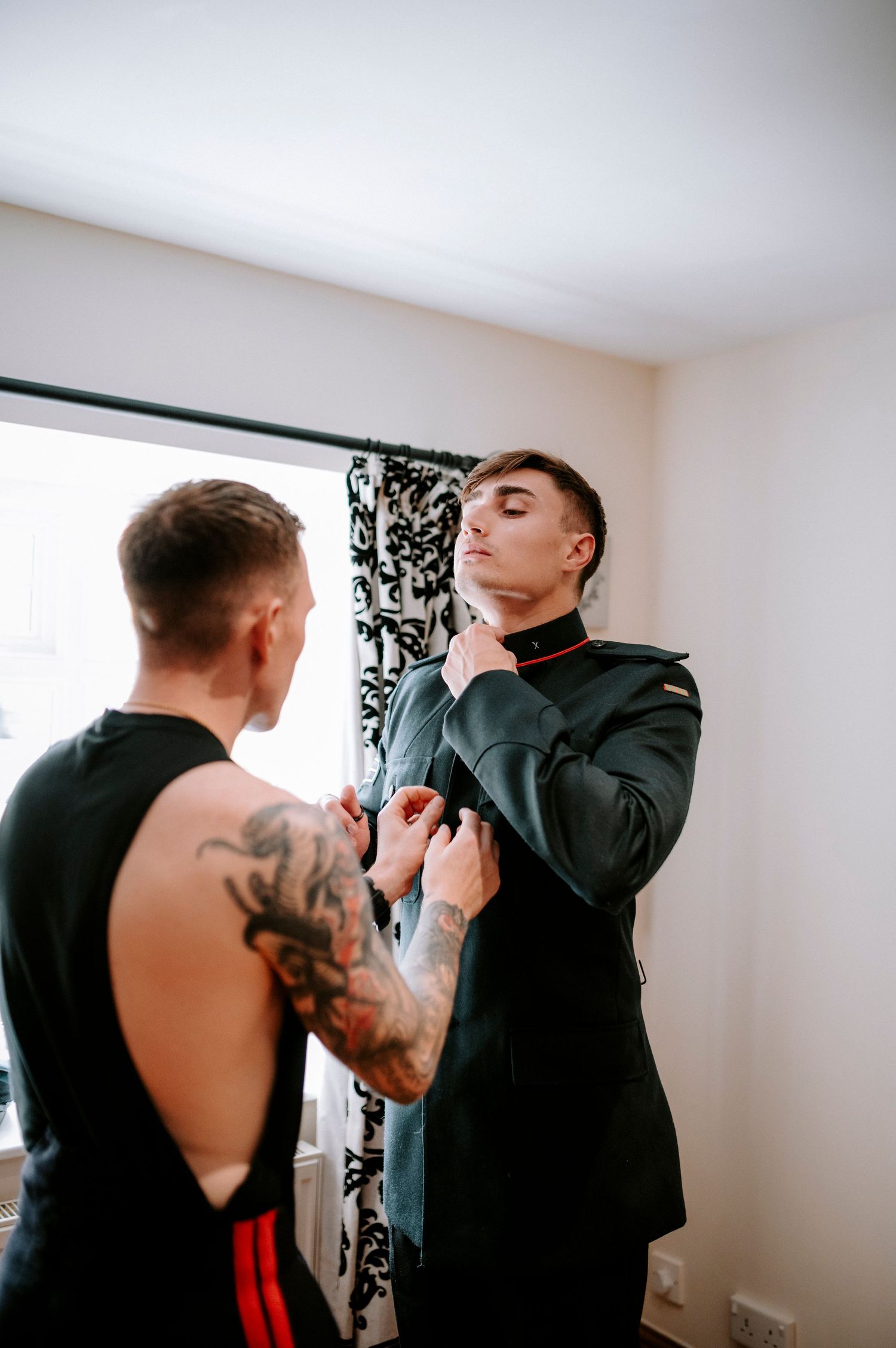 Two people helping with formal attire preparations in a bright room.