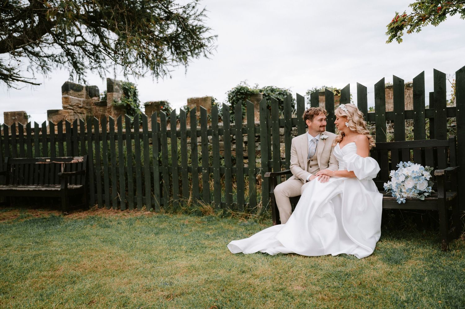 Newlyweds sitting together on grass by rustic wooden fence during outdoor wedding celebration.