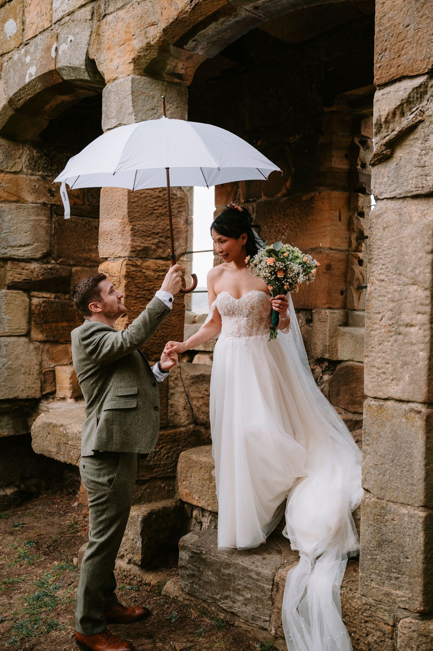 Wedding couple poses under stone arches with white umbrellas and floral bouquet.