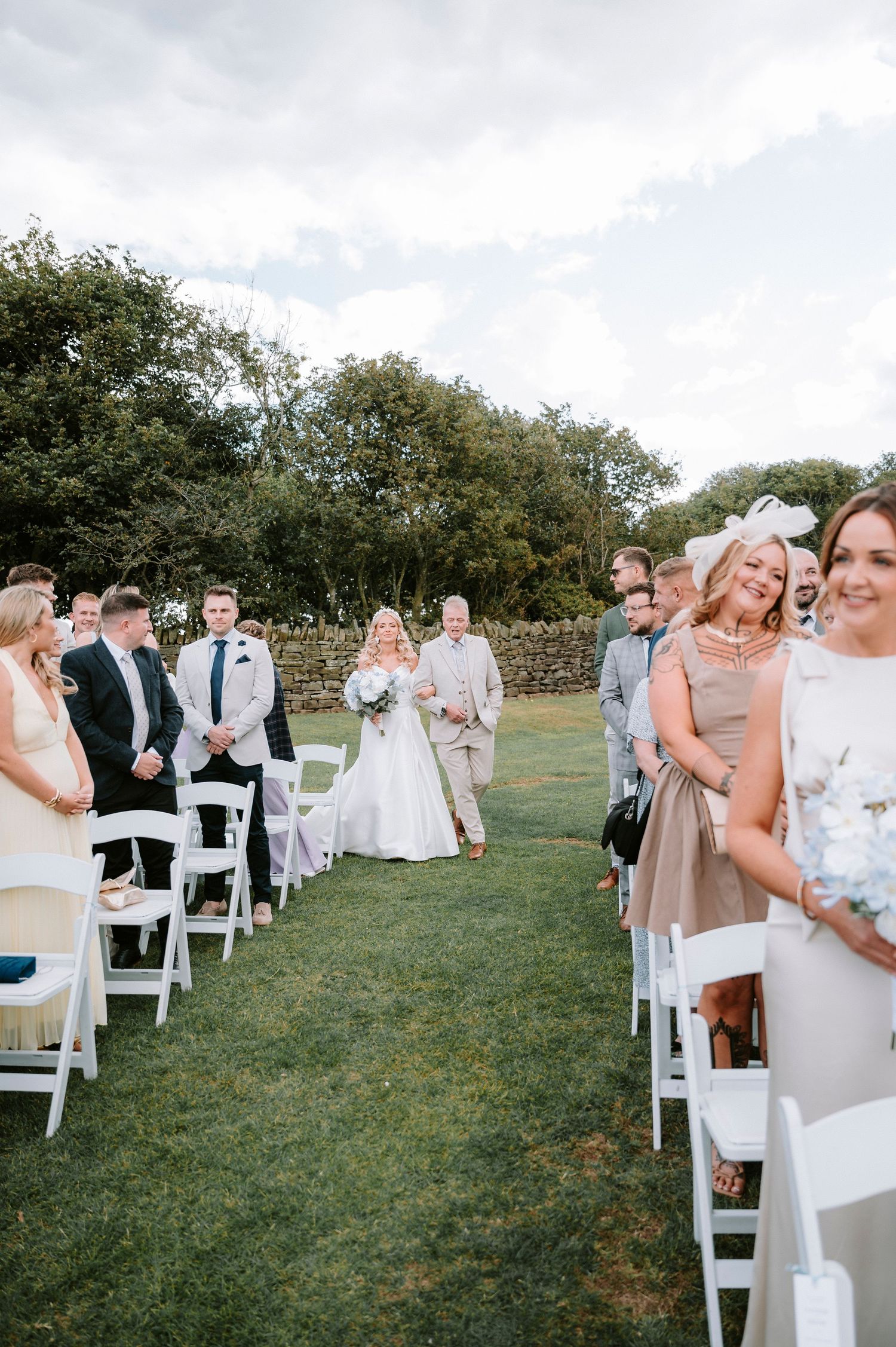 Wedding guests seated in white chairs on a grassy lawn for an outdoor ceremony with stone wall backdrop.