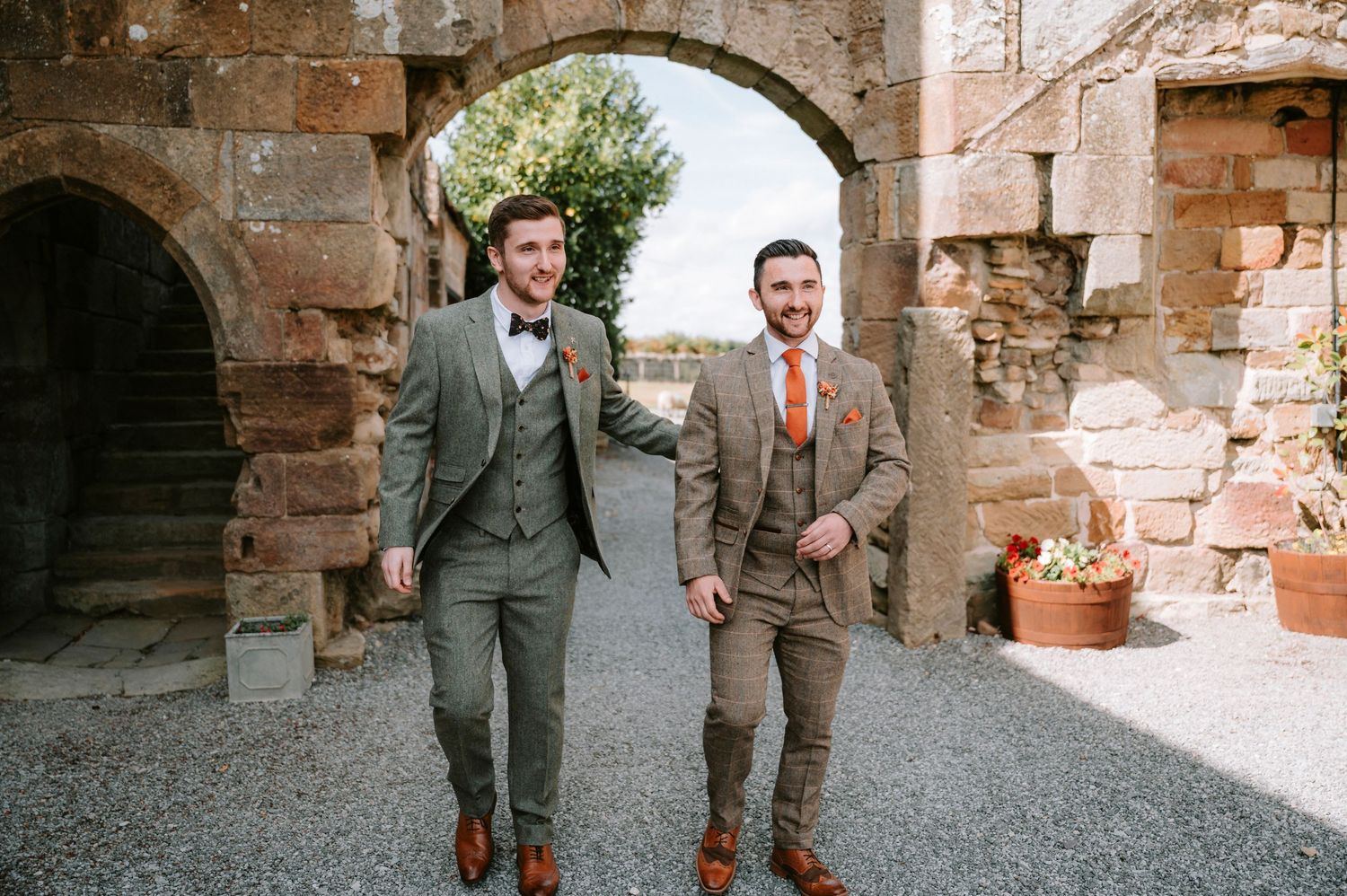 Groomsmen in grey suits walking through stone archways at a rustic venue.