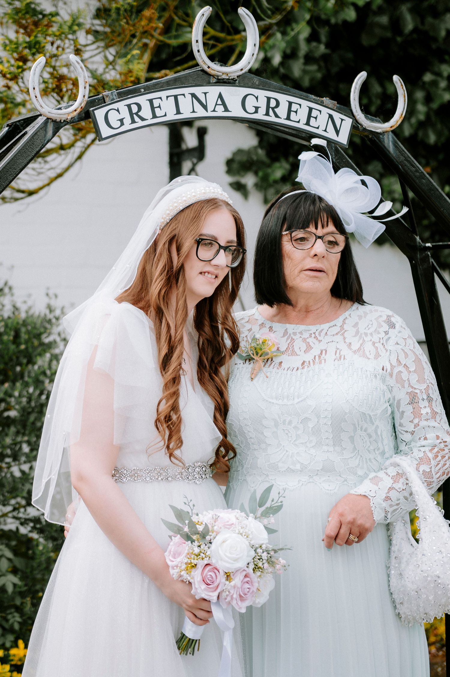 Two people in white dresses standing under a Gretna Green archway with horseshoes.