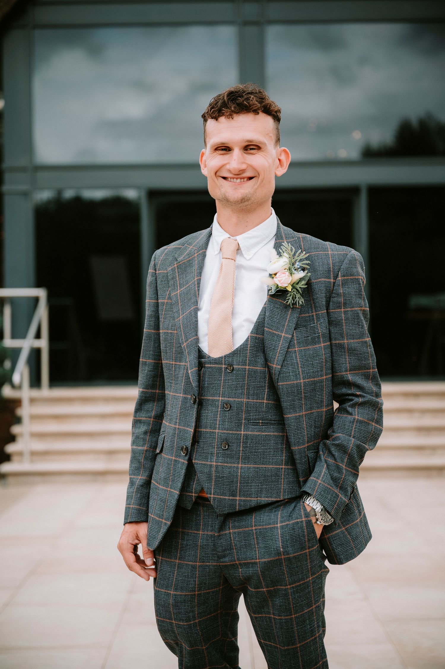 Groom in grey plaid three-piece suit stands confidently outside modern building.