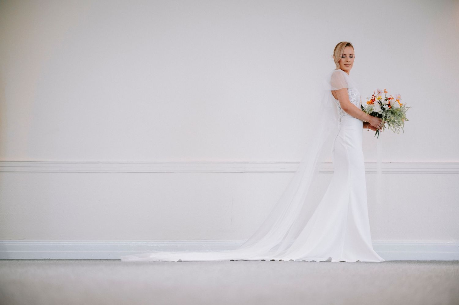Minimalist wedding portraits against a white wall showcase a flowing wedding dress and veil.