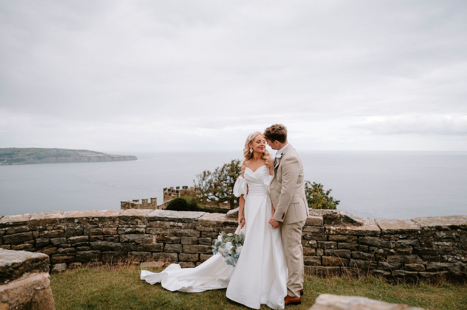 Couple in wedding attire embraces on a scenic coastal cliff with dramatic cloudy skies in the background.