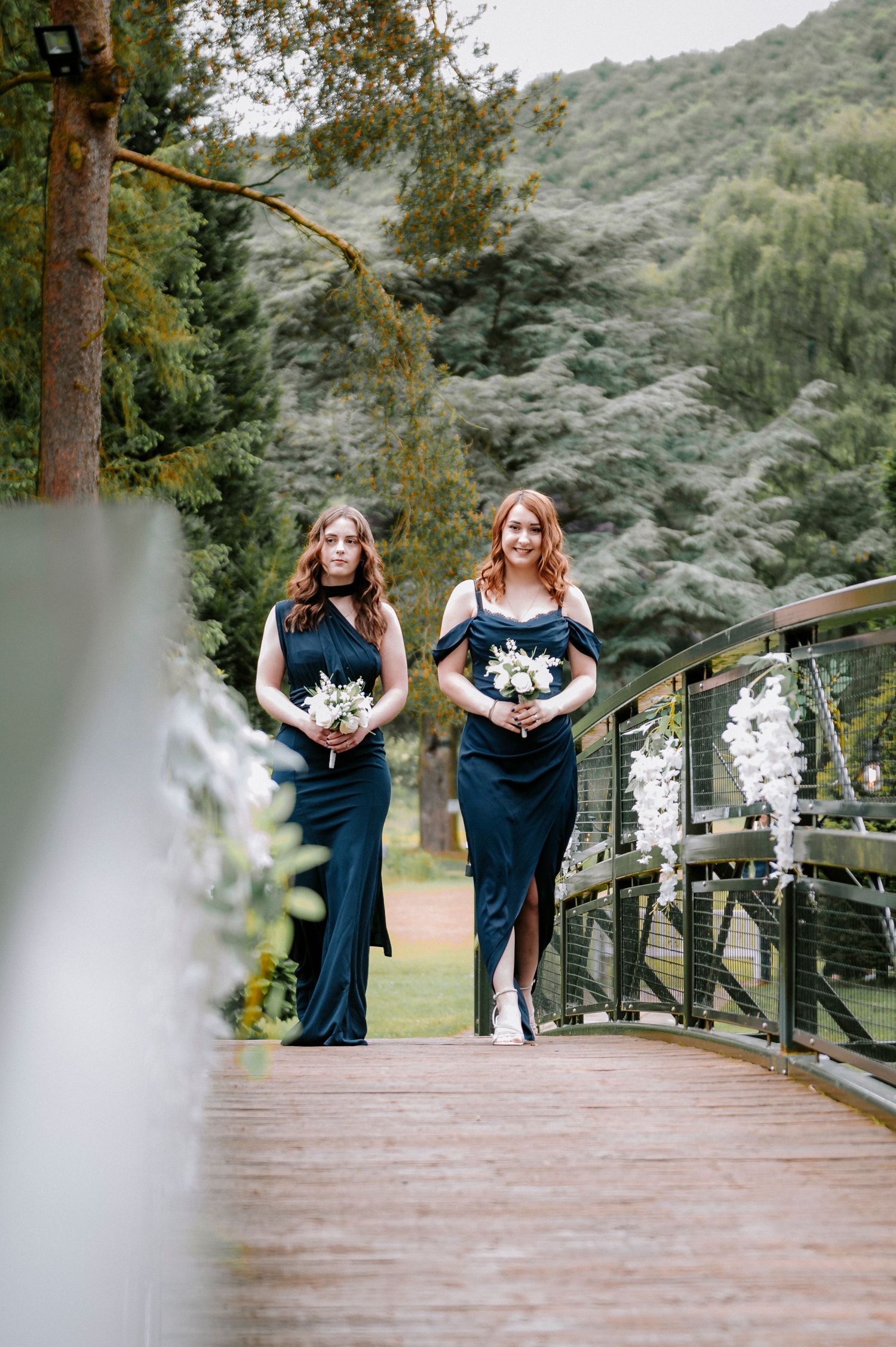 Bridesmaids in navy blue dresses walking across a wooden bridge decorated with white flowers.