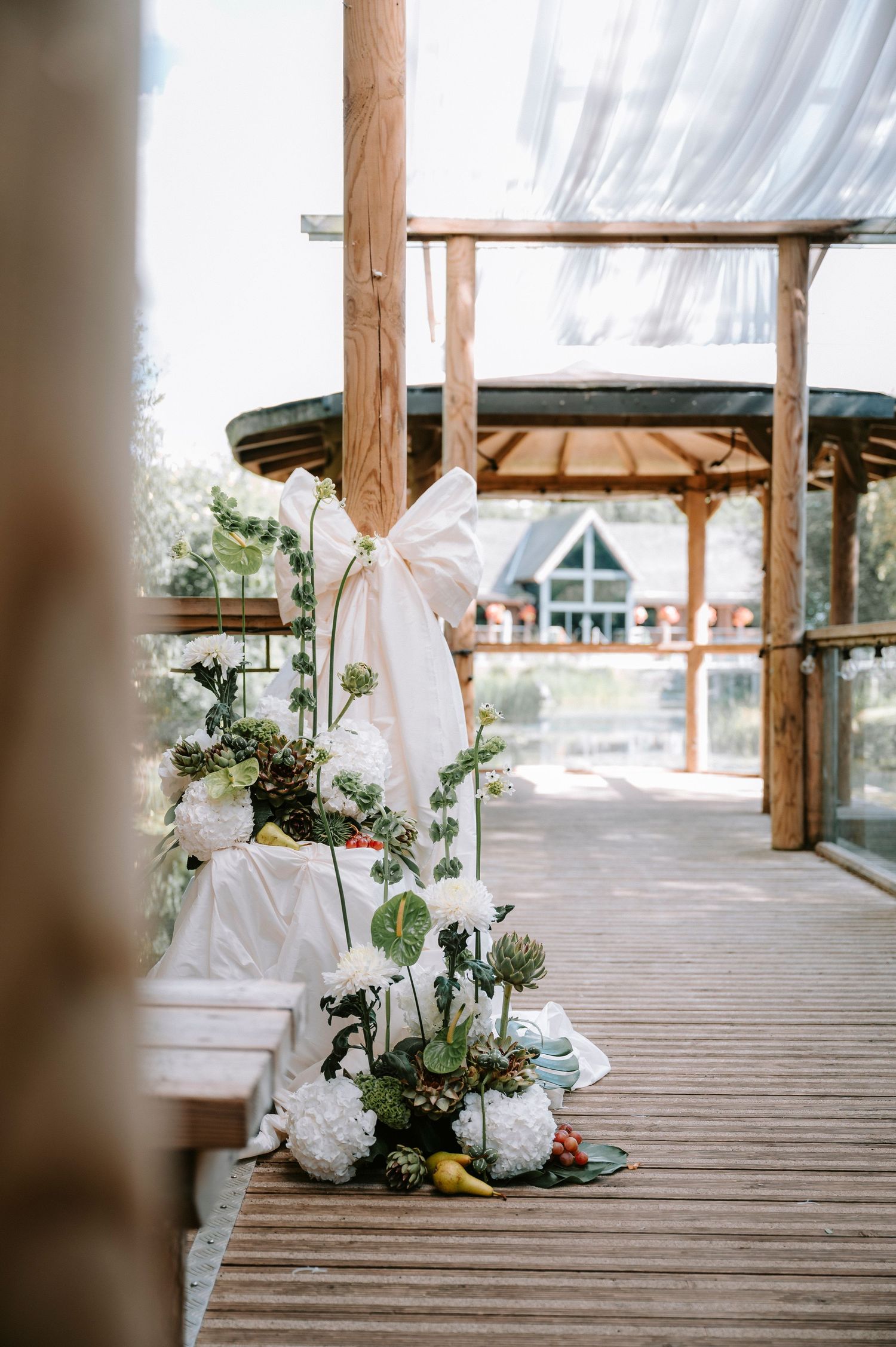 Elegant white floral arrangements with greenery decorate a wooden deck at an outdoor wedding venue.