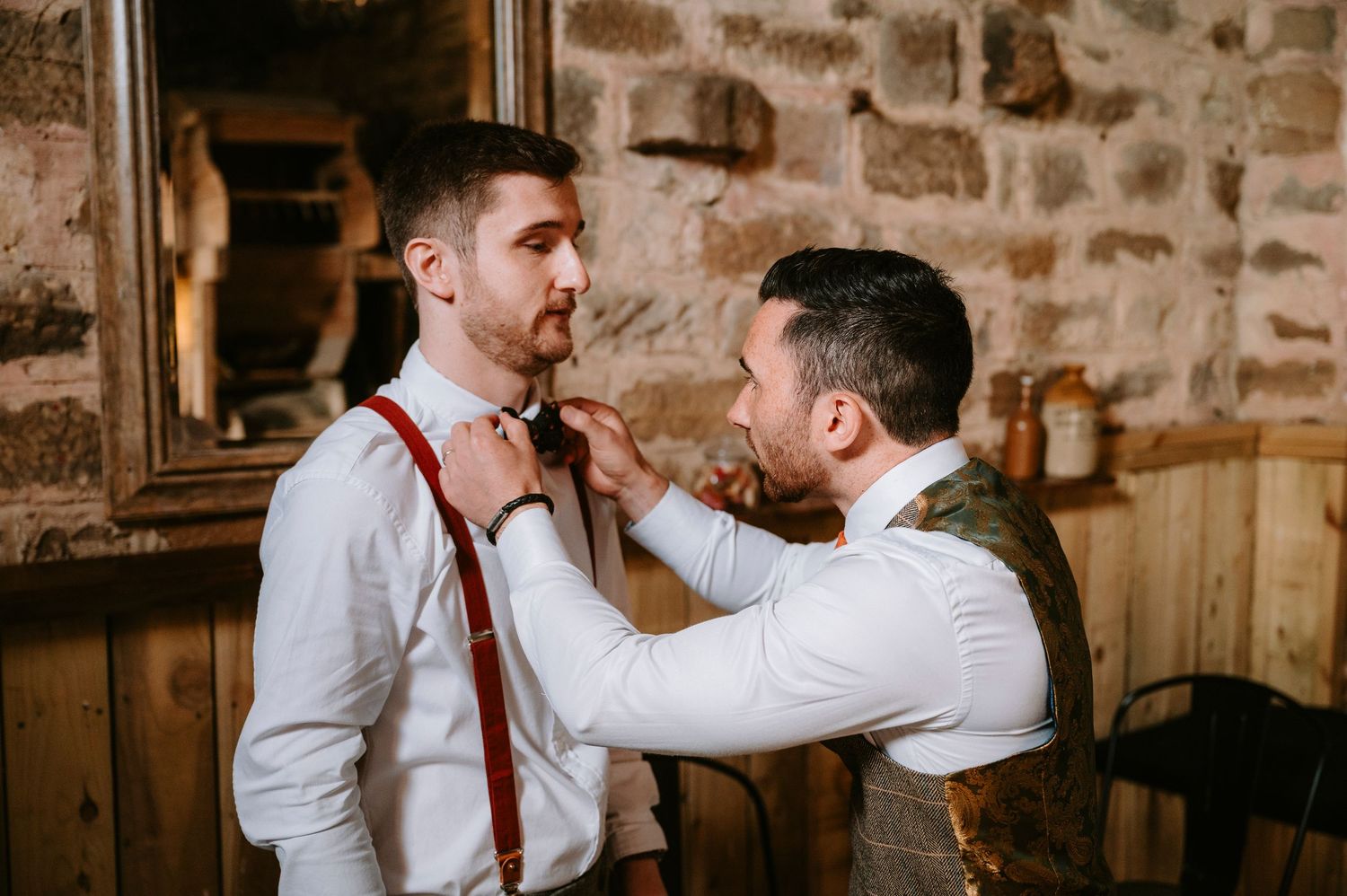 Groomsmen adjusting ties in rustic stone-walled room.