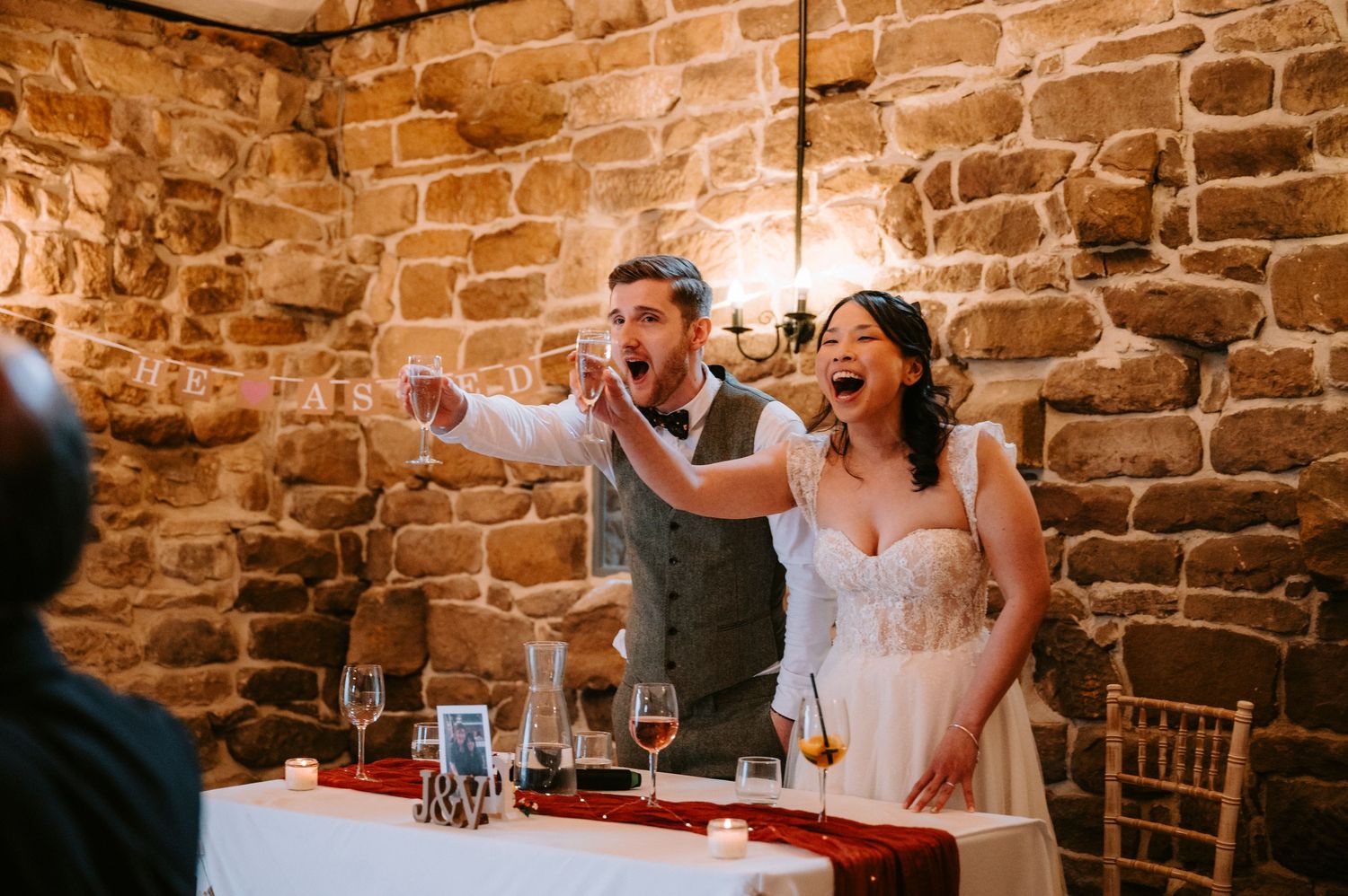 Wedding reception celebration in a rustic stone-walled venue with warm lighting and elegant table settings.