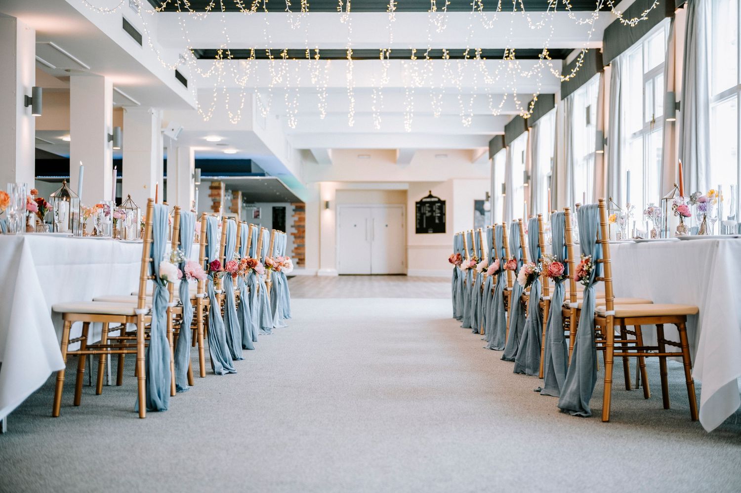 Long banquet tables set with elegant place settings in a bright, airy venue with tall windows.