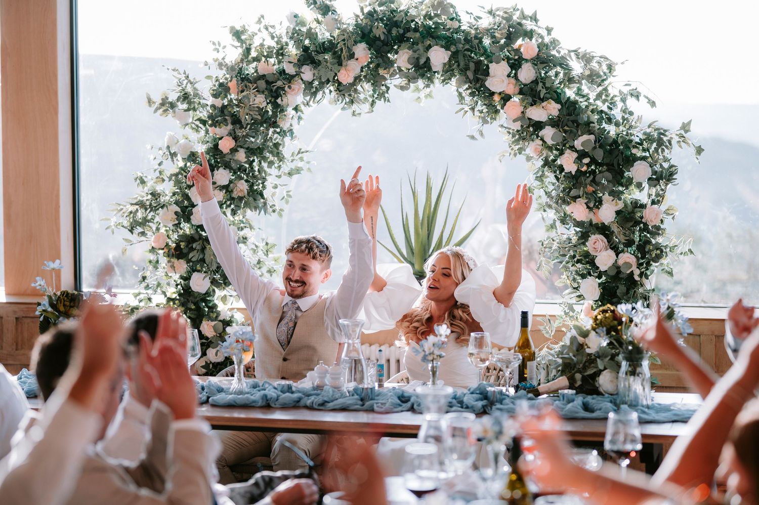 Joyful celebration at an outdoor wedding reception under a floral arch with pink and white roses.