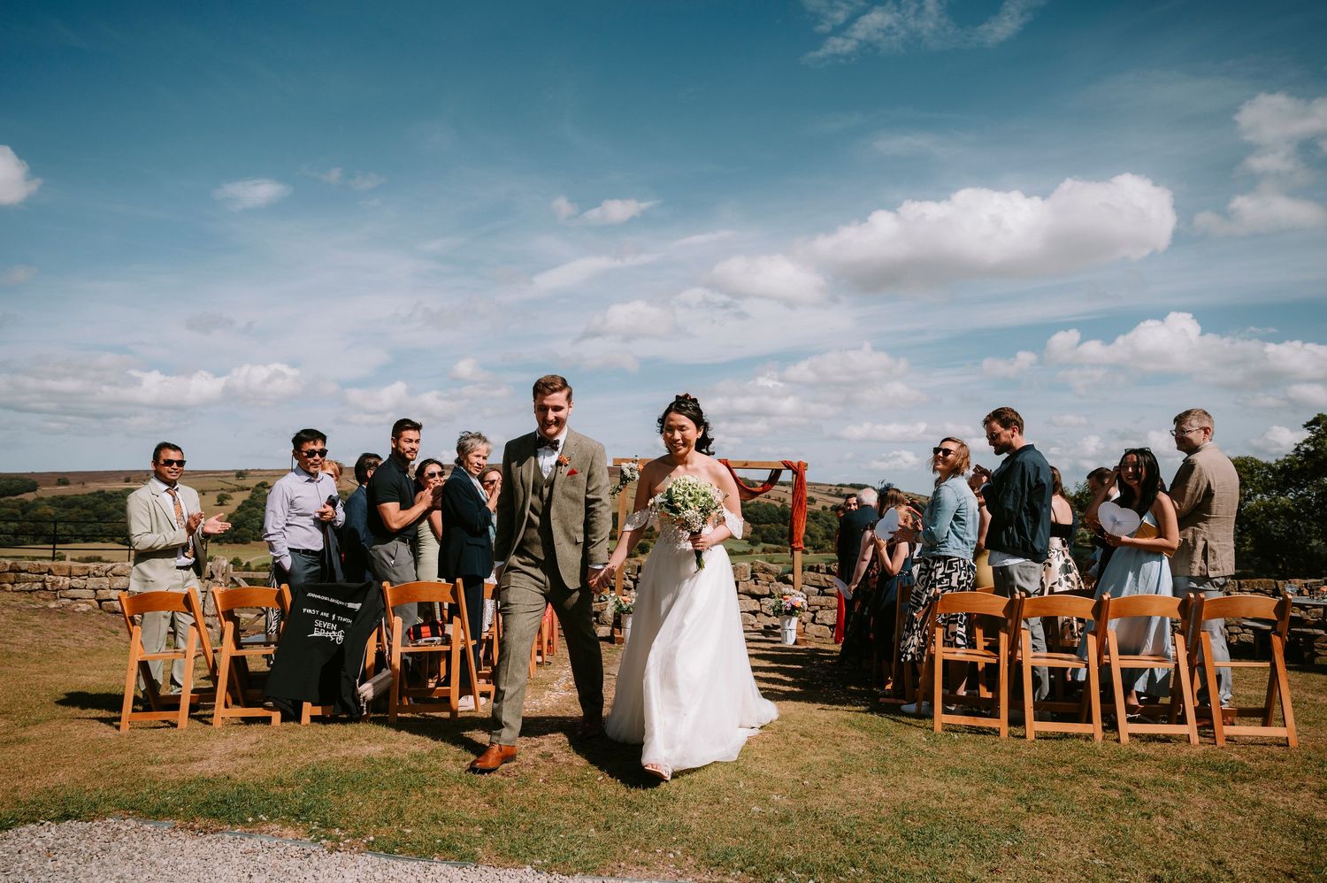 Wedding party and guests gathered for outdoor ceremony against blue sky and rolling hills.