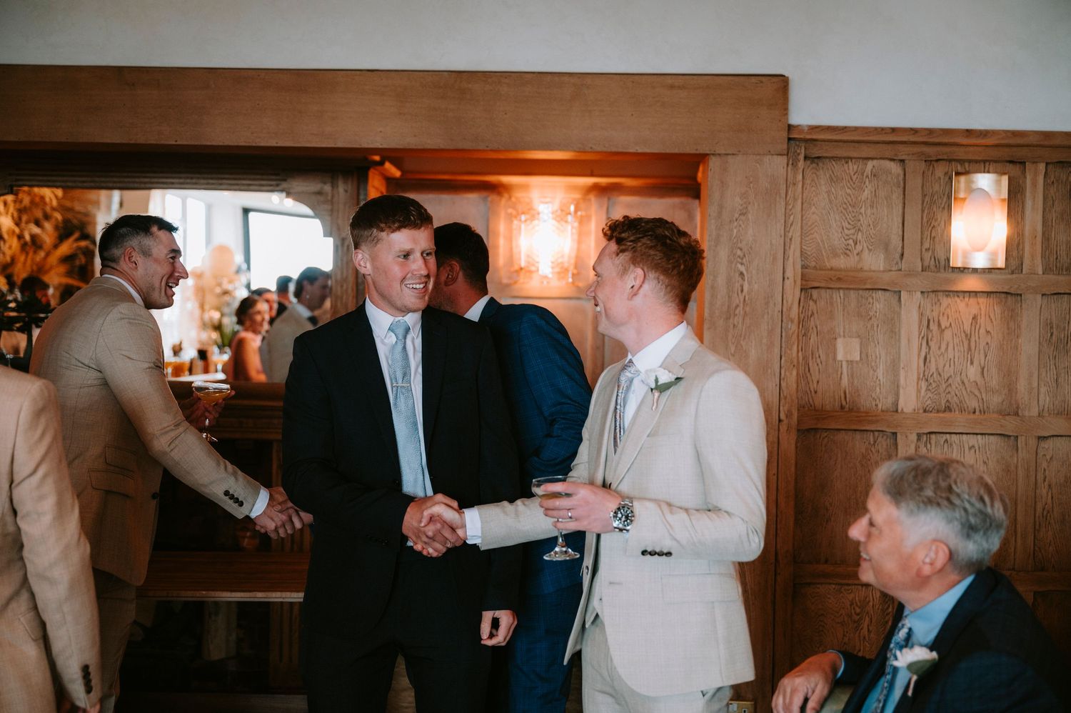 Two people shake hands during a wedding reception in a rustic wooden venue with warm lighting.