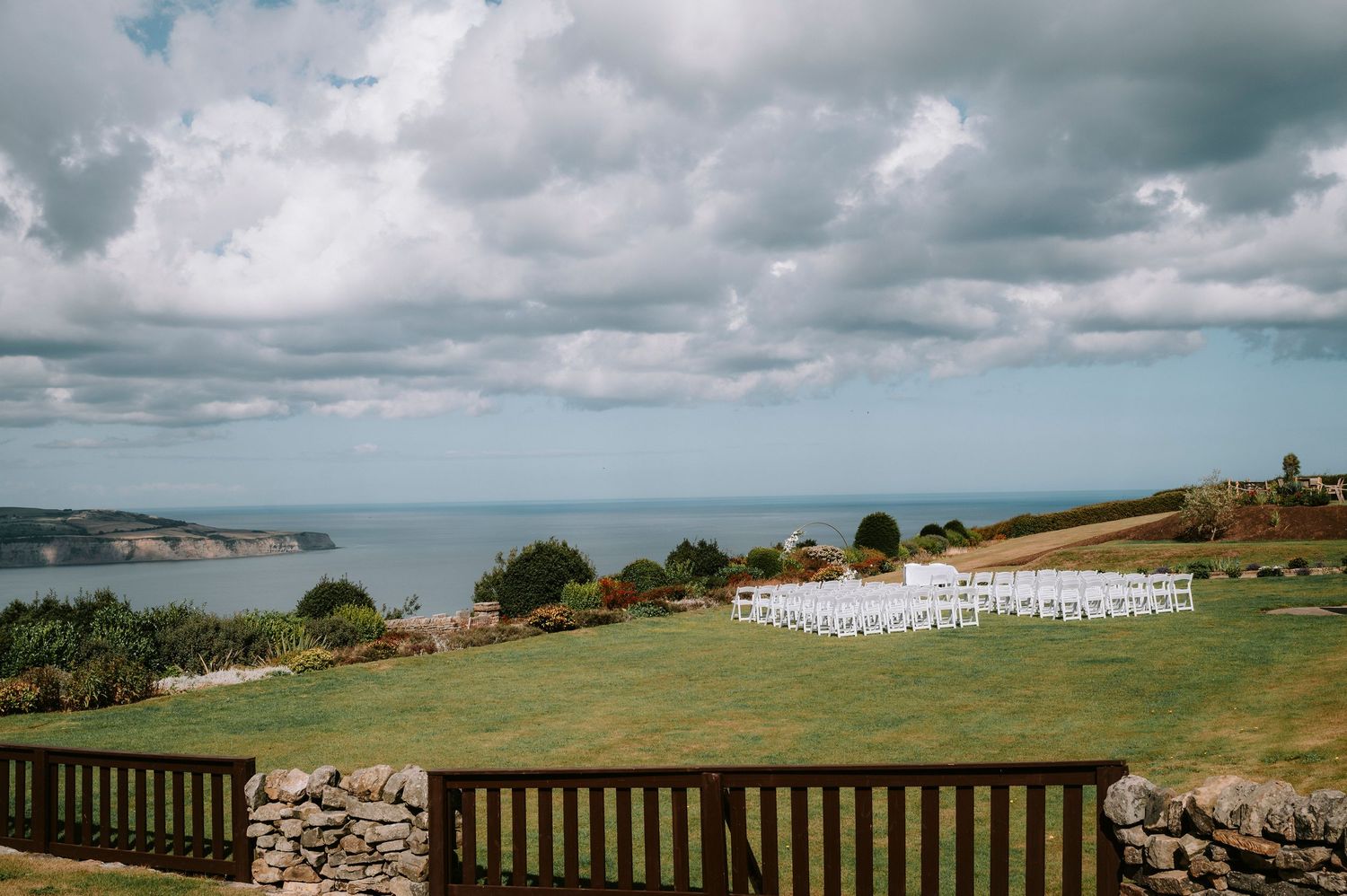 Coastal wedding ceremony setup with white chairs overlooking ocean view under cloudy sky.