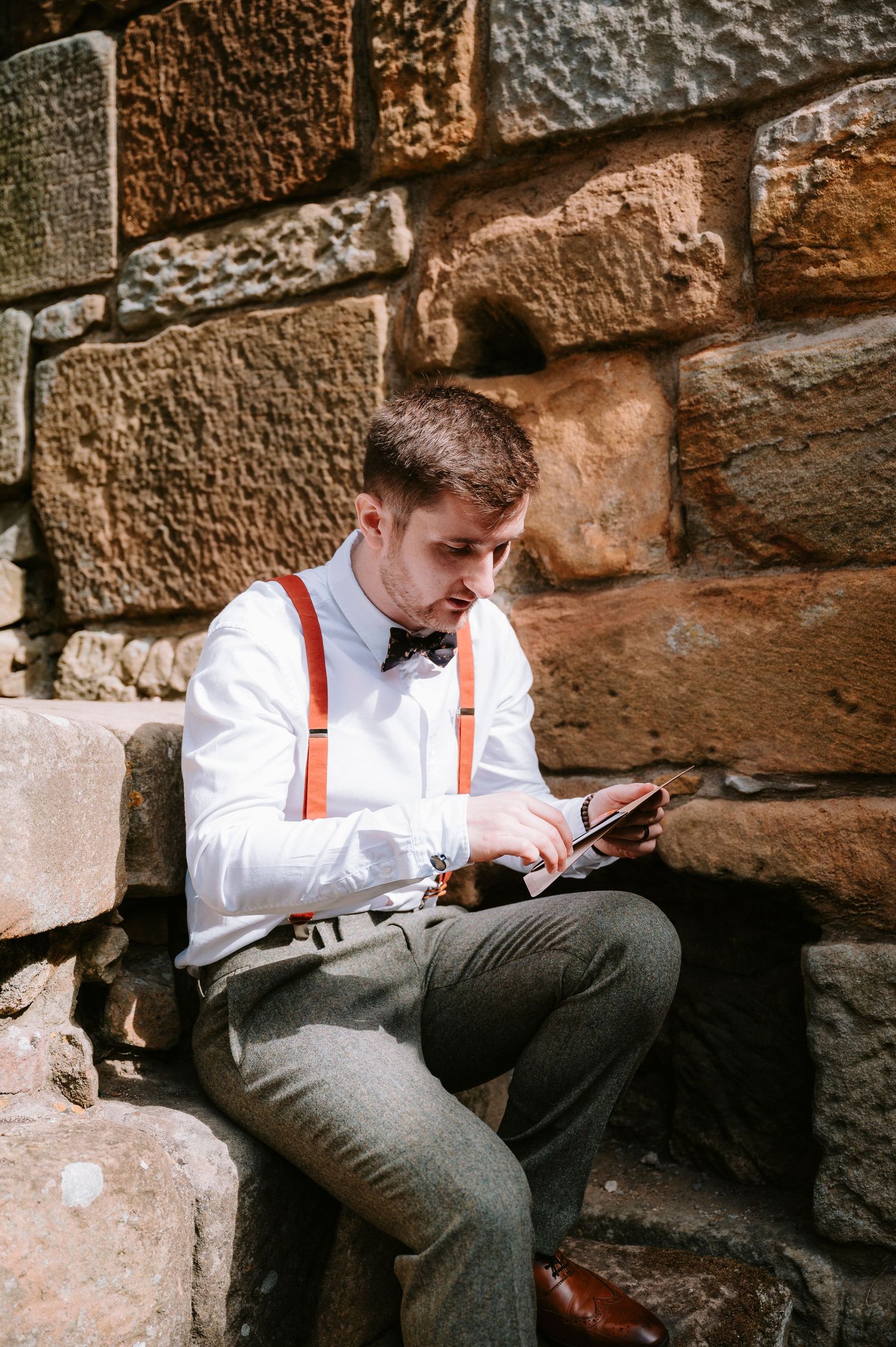 Person in white shirt and suspenders sitting against stone wall looking at mobile phone.