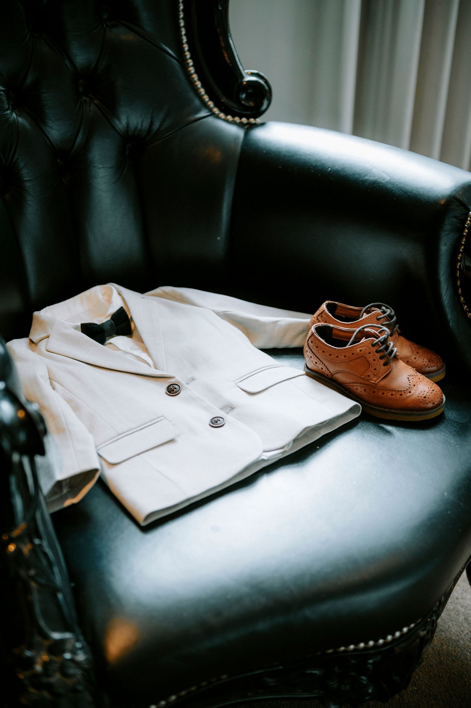 Wedding attire including a white shirt and brown leather shoes arranged on a black leather couch.