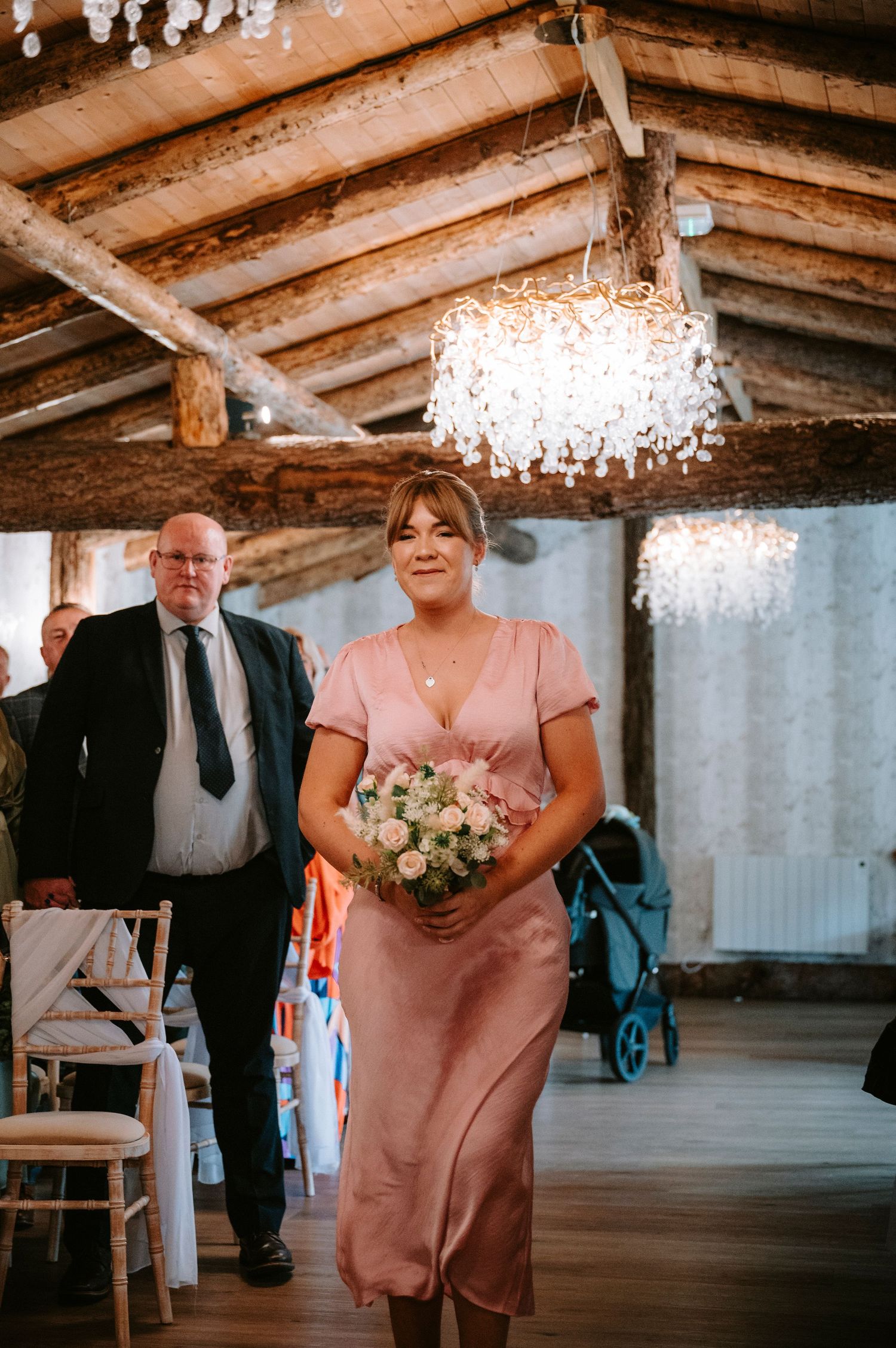 Wedding party member in blush pink dress carries bouquet down rustic aisle under crystal chandelier.