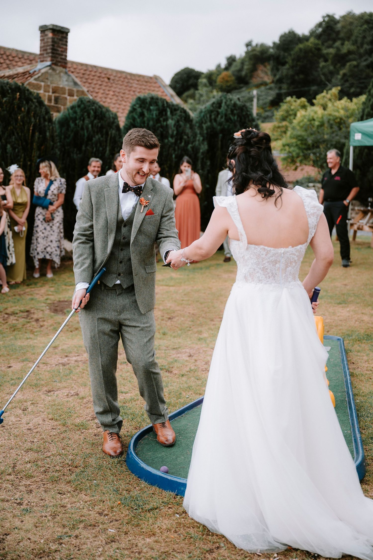 Newlyweds play mini golf together on their wedding day as guests watch and smile.